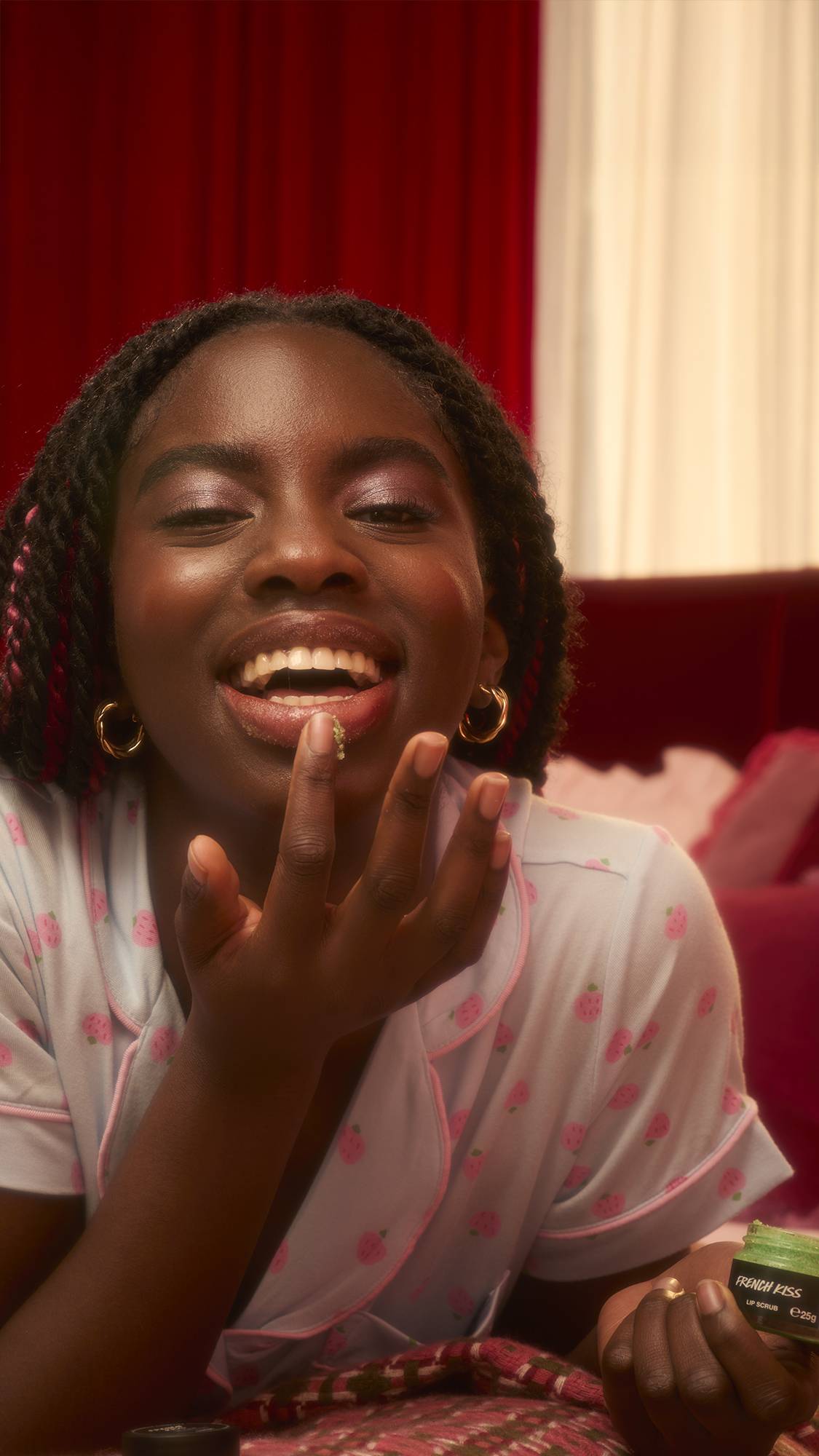 A woman in pink strawberry pyjamas laughs while applying green "French Kiss" lip scrub to her bottom lip. She holds the open jar in her other hand while reclining in a warm, dimly lit room with red curtains.
