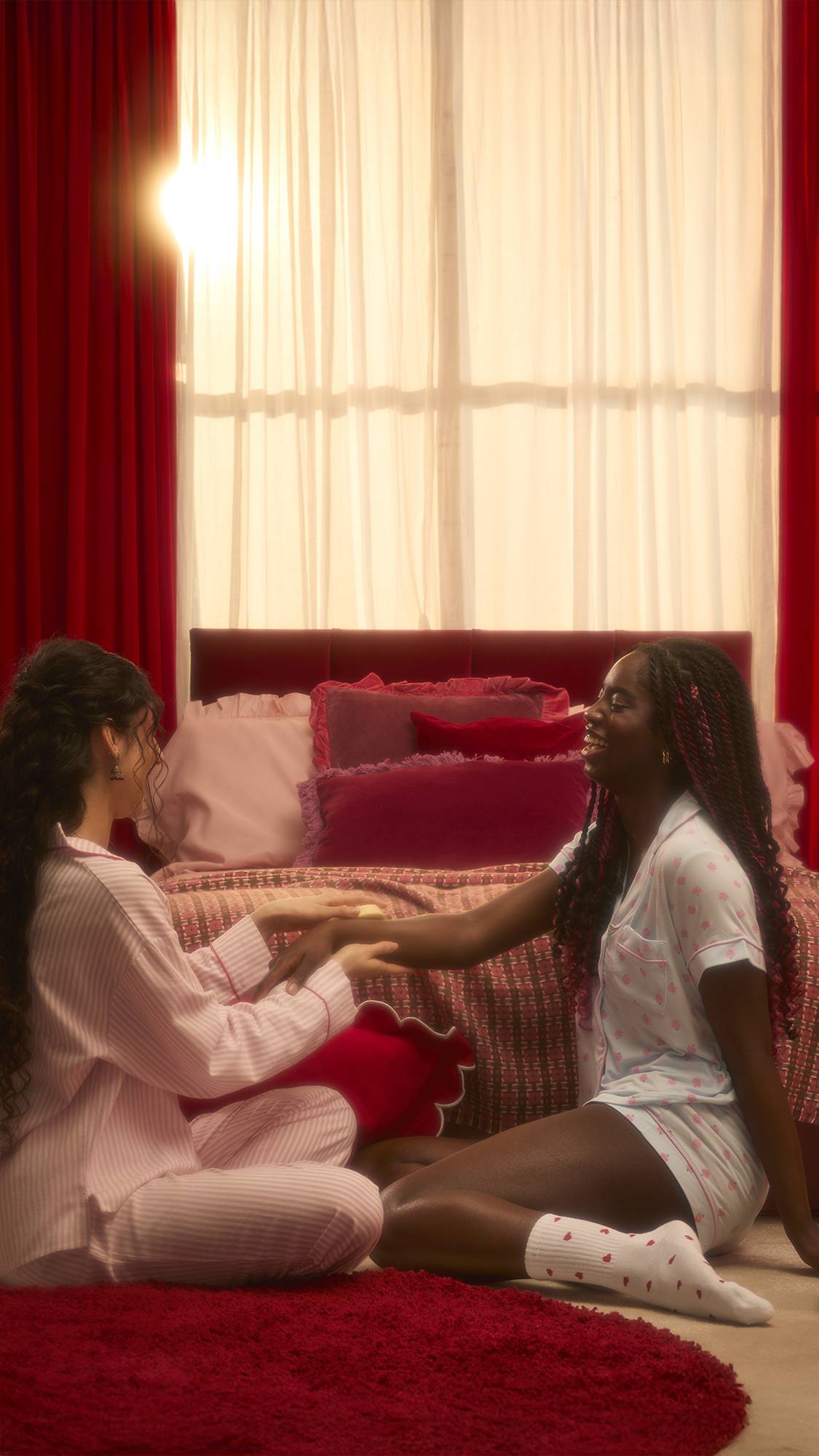 Two people sit in a sunlit bedroom with red curtains, wearing strawberry-patterned and striped pyjamas. One person gently massages the other's arm with a cream-coloured Gorgeous massage bar.