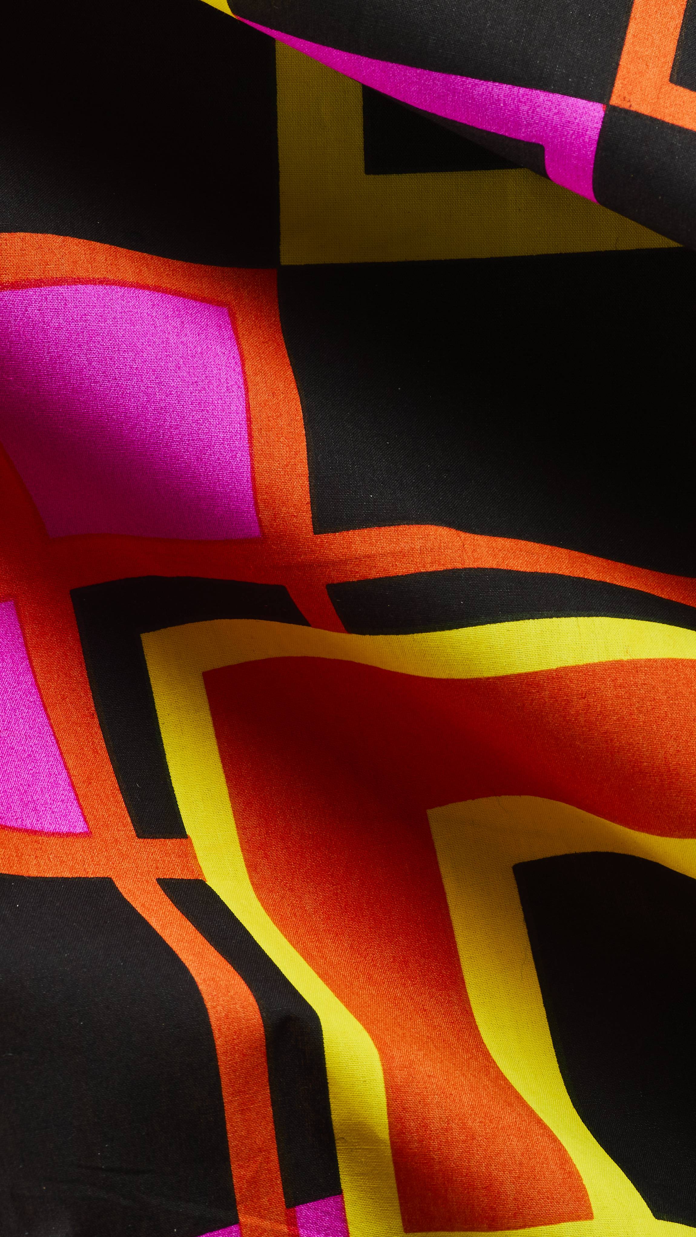 A close-up of a draped LUSH fabric knot wrap featuring a bold geometric pattern of interlocking squares and lines in vivid yellow, orange, magenta pink, and black.