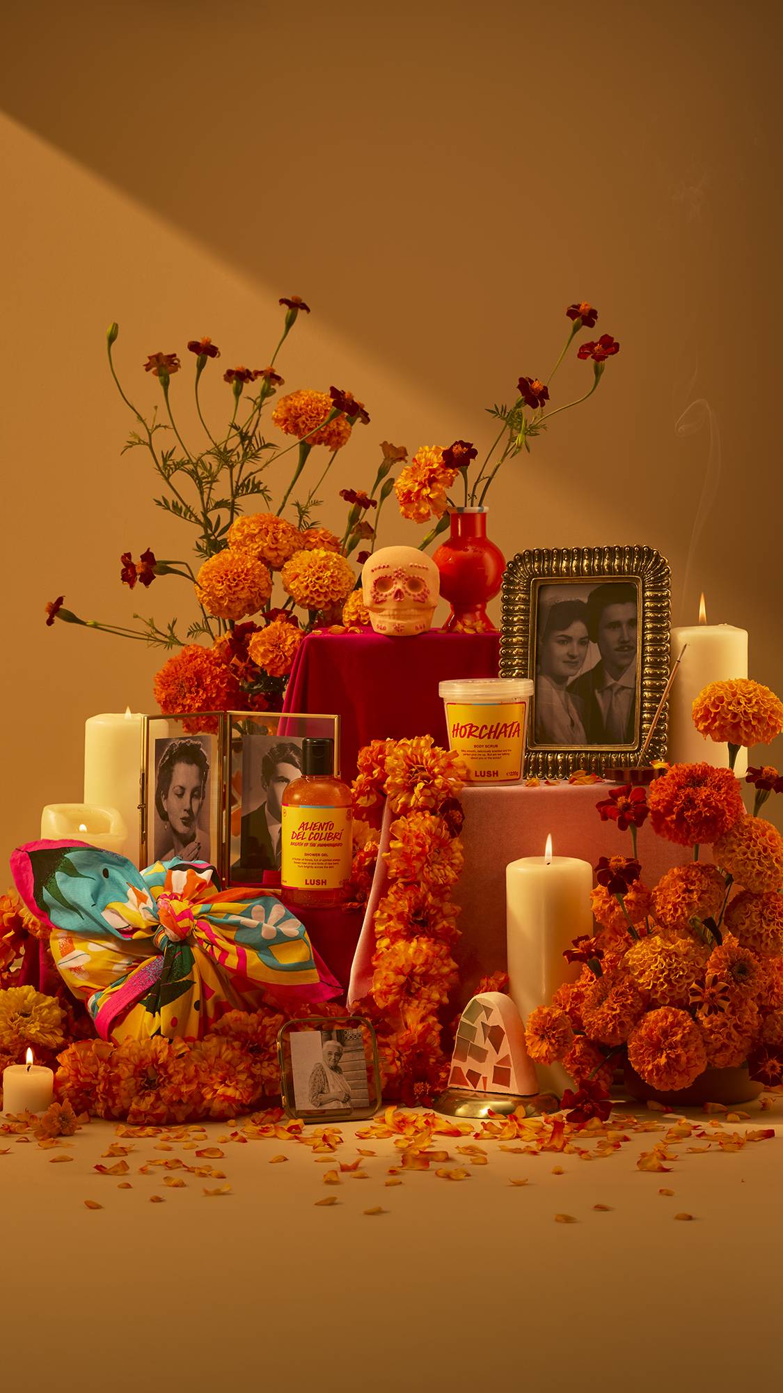 A Día de Muertos altar arrangement featuring orange marigolds, lit candles, framed photos, with the range of Lush products, including a skull bath bomb, shower gel, body scrub, soap, and a colourful knot-wrap gift.