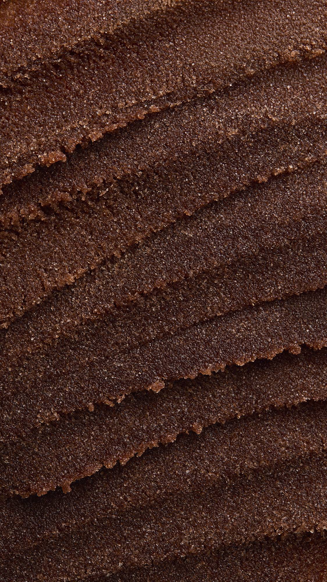 A macro close-up texture shot of rich, dark brown sugar lip scrub spread in thick, textured diagonal ridges across the frame with fine sugar crystals visible throughout.