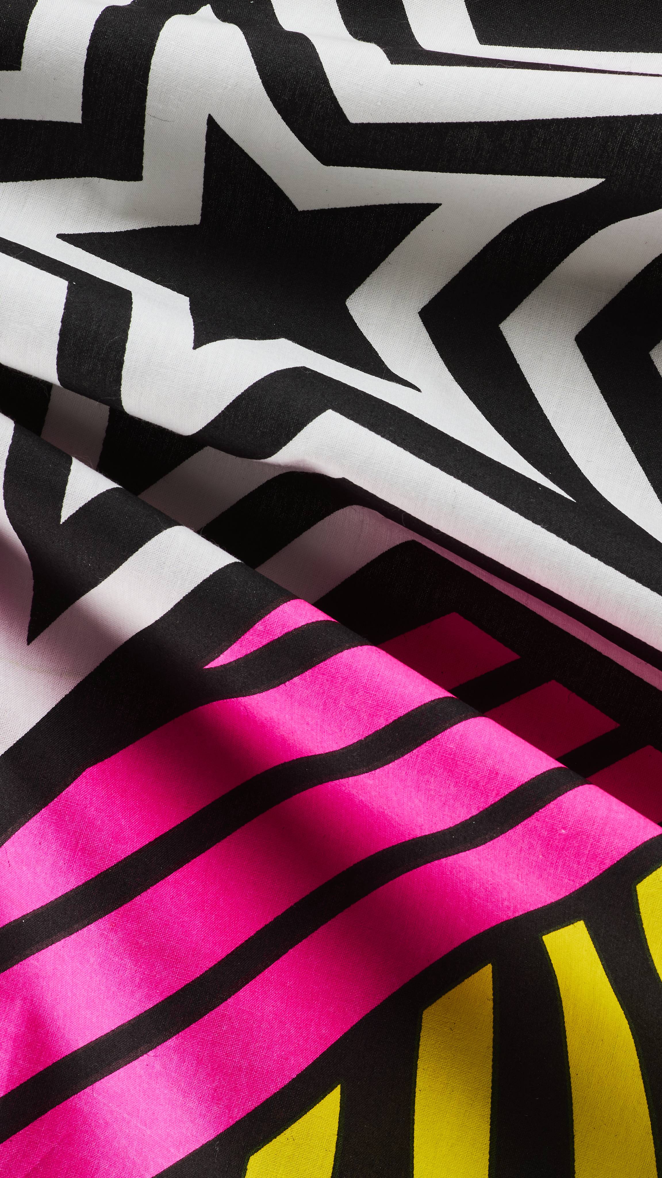 A close-up of draped fabric knot wrap highlighting a bold graphic design with black and white repeating star patterns, vivid neon pink stripes, and bright yellow accents.