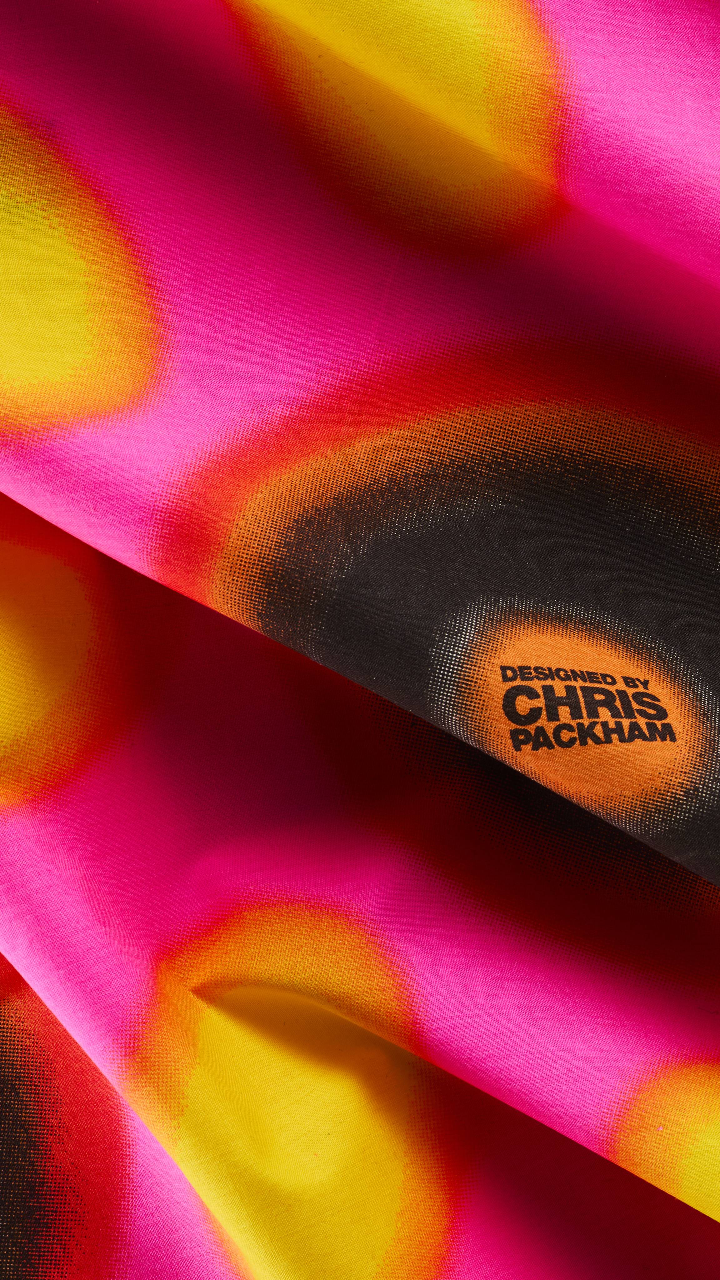 A close-up of draped fabric knot wrap featuring a tie-dye pattern in vibrant pink, orange, yellow, and black, detailed with a small circular stamp that reads "DESIGNED BY CHRIS PACKHAM".
