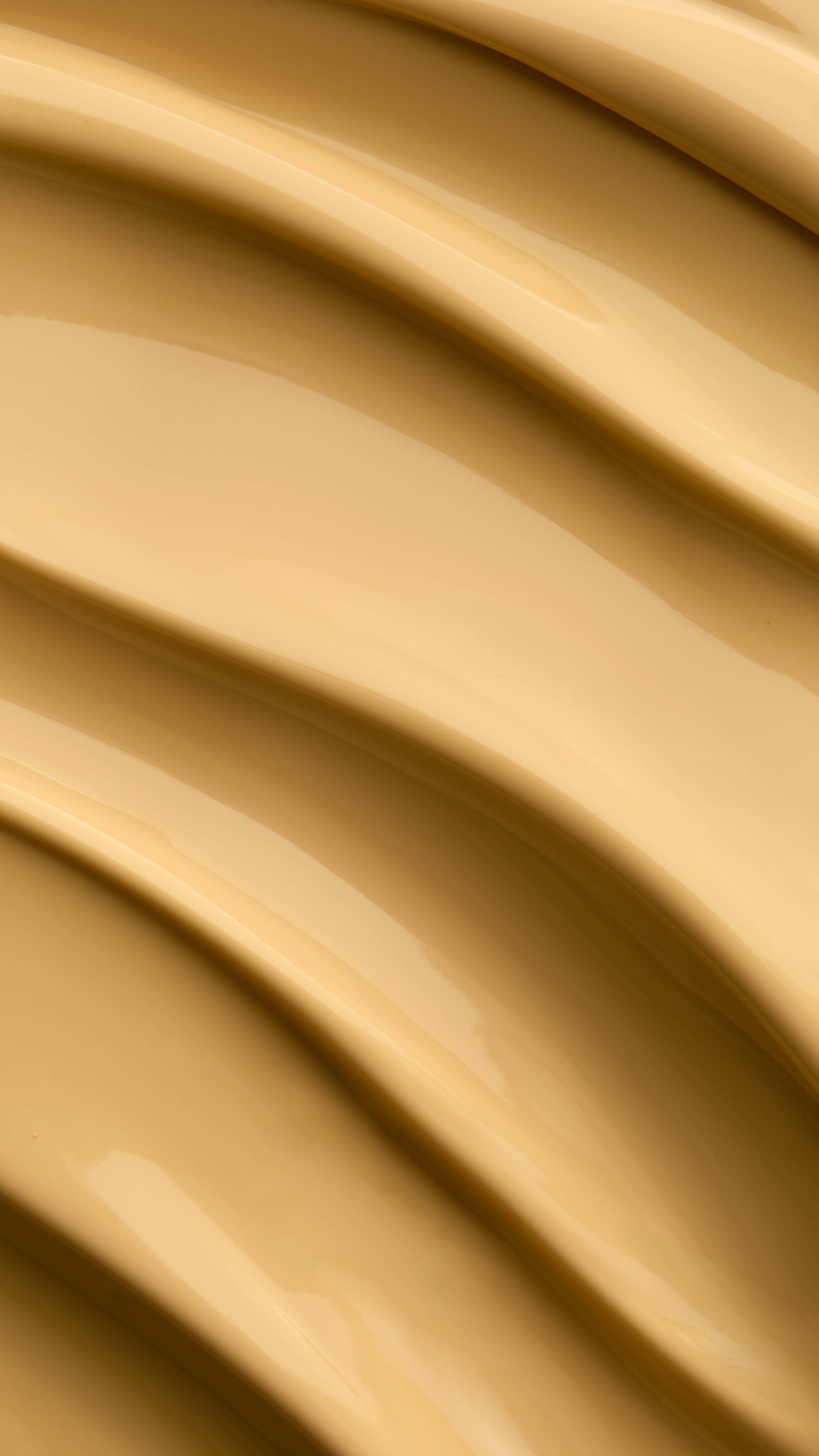 A macro texture close-up showing smooth, silky waves of rich, warm yellow "Mango Kulfi" body lotion with glossy highlights and subtle shading.