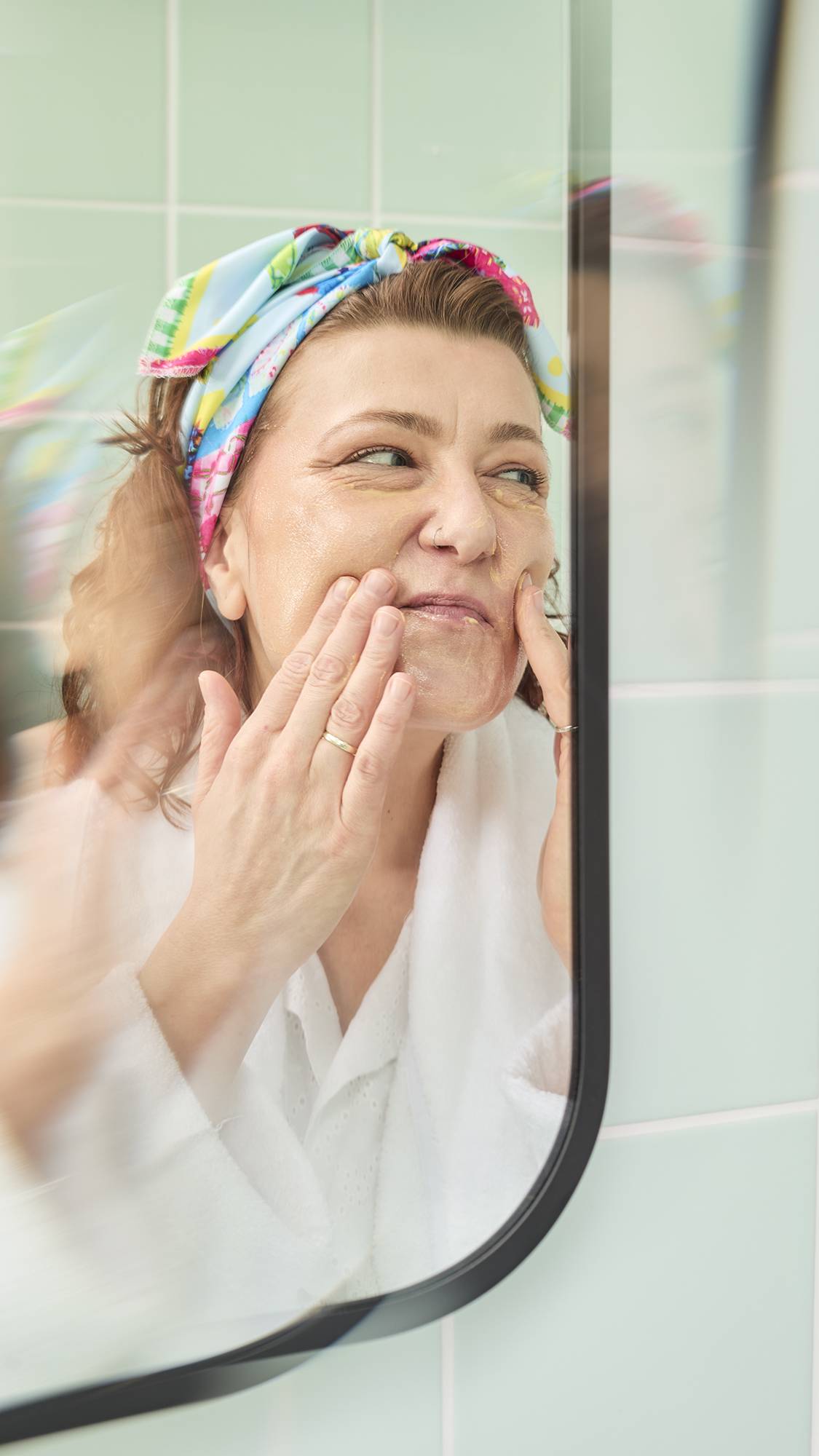 The image shows a model with wavy auburn hair in a bathroom mirror, smiling while applying a foundation with her hands. She has fashioned the colourful "My Family Houses" fabric knot wrap into a stylish headband.