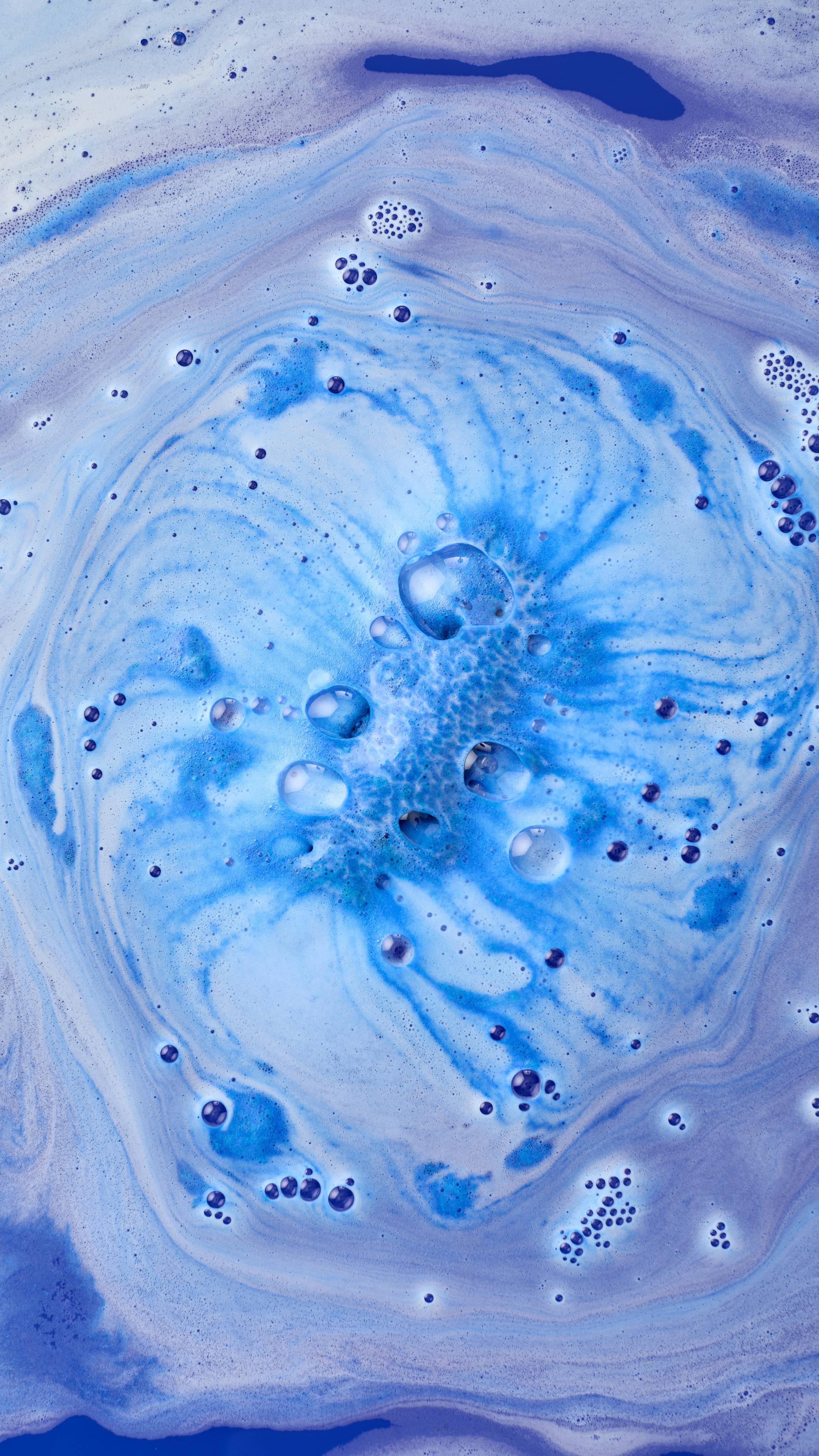 An aerial view of a vibrant blue "Peace" bath bomb dissolving in water. It creates a swirling vortex of purple, blue and white foam, with small bubbles radiating outwards from the centre.