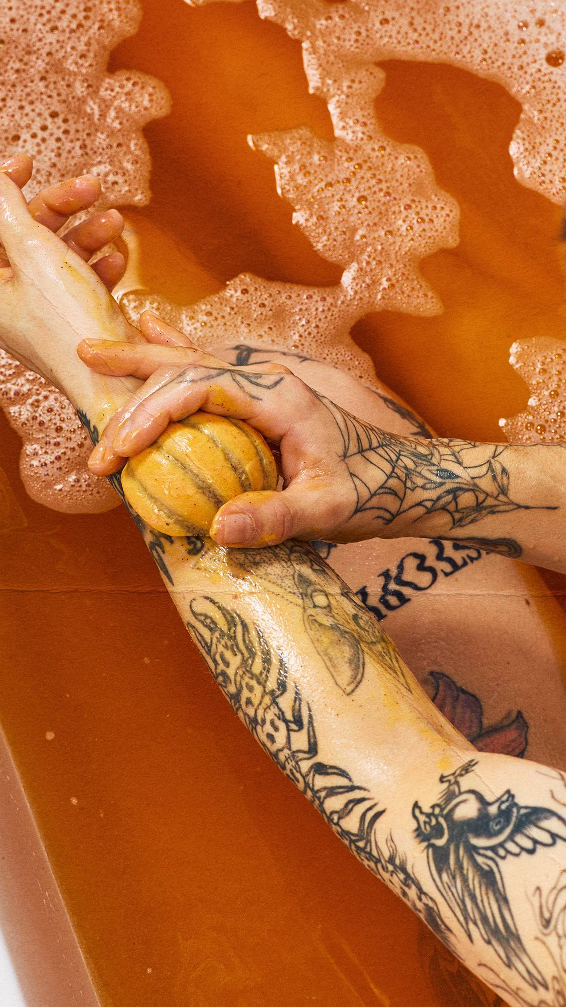 Tattooed hands rub an orange, pumpkin-shaped solid body scrub over a wet forearm above warm, amber bathwater filled with floating, foamy bubbles.