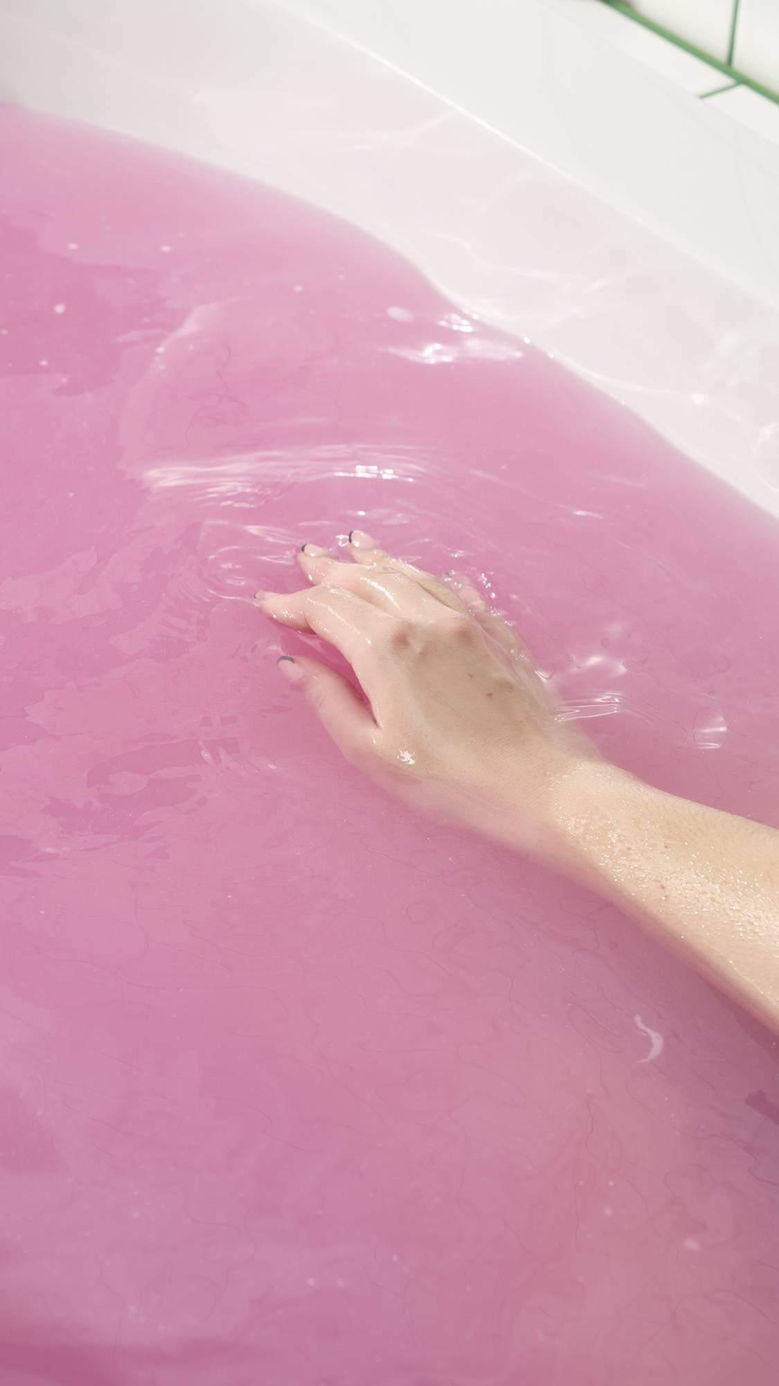 The Sex Bomb Bath Bomb has completely dissolved, leaving the bathtub filled with a vibrant, opaque shade of deep pink. A hand is shown gently submerged in the colorful water, illustrating the relaxing and aesthetically pleasing result of the bath.