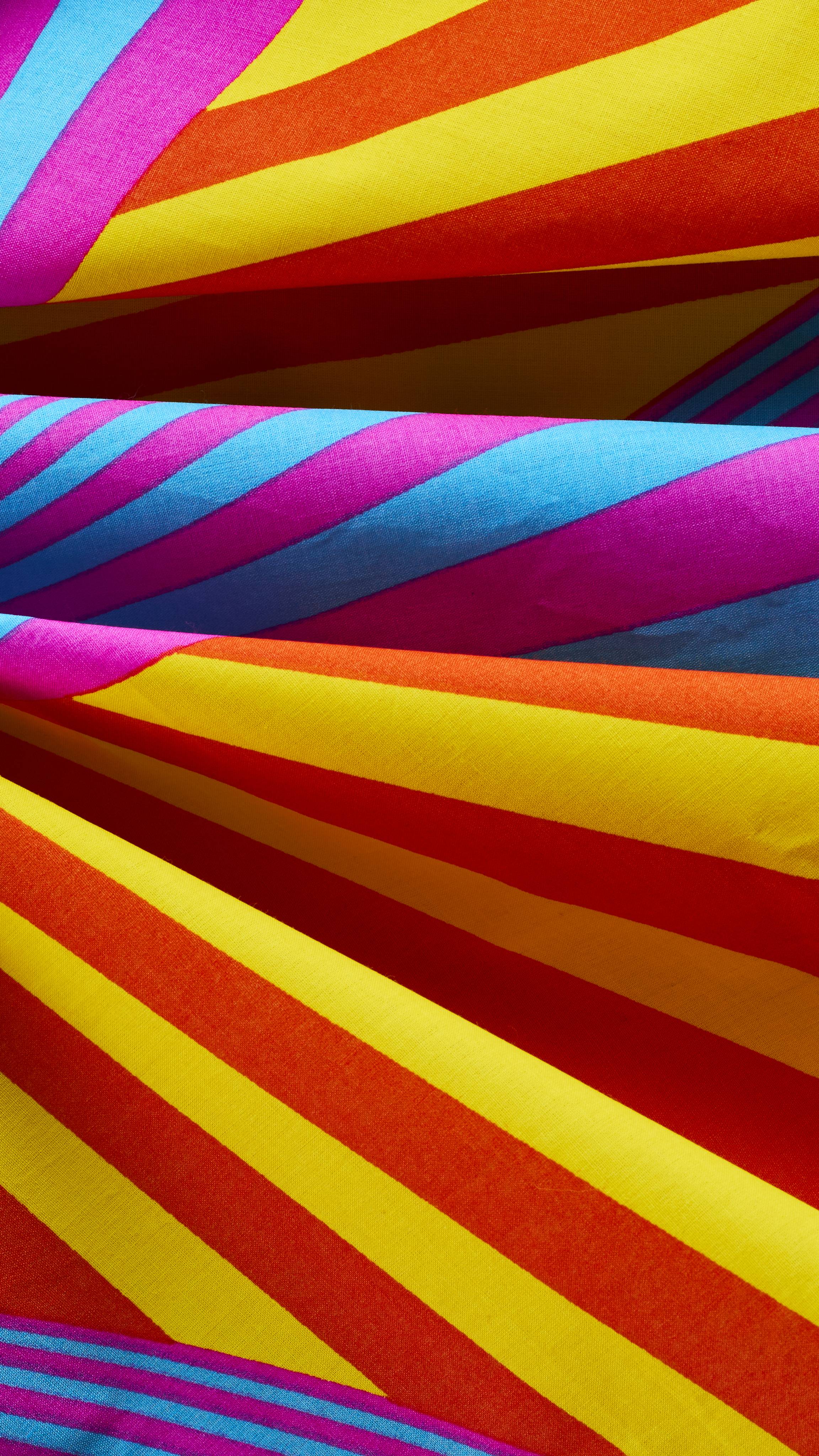 A close-up of a vibrant knot-wrap fabric showing draped folds of bold, diagonal stripes in bright yellow, orange, magenta, and sky blue.