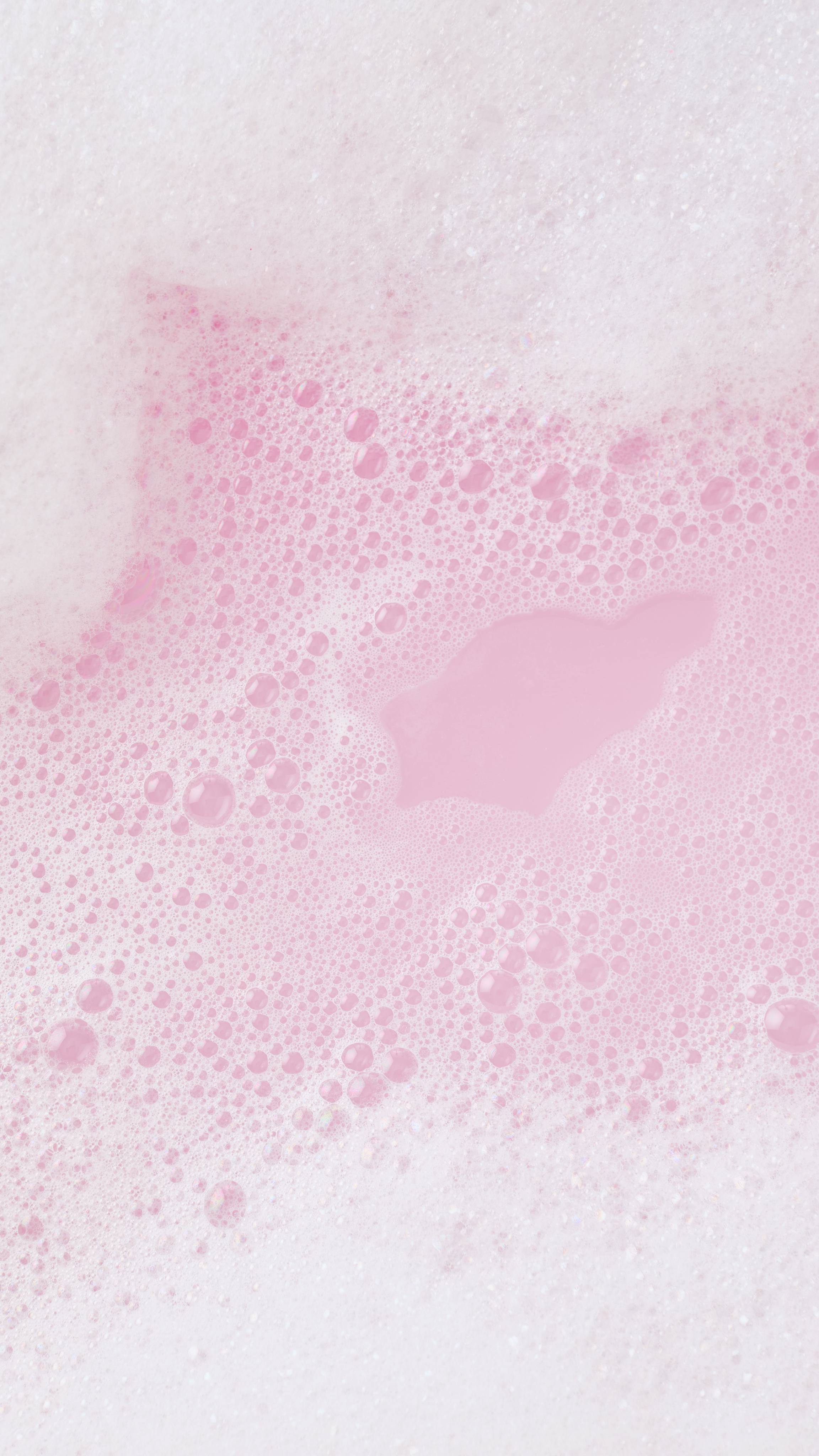 A thick layer of soft white bath foam parts to reveal pastel pink bathwater beneath, surrounded by clusters of fine frothy bubbles.