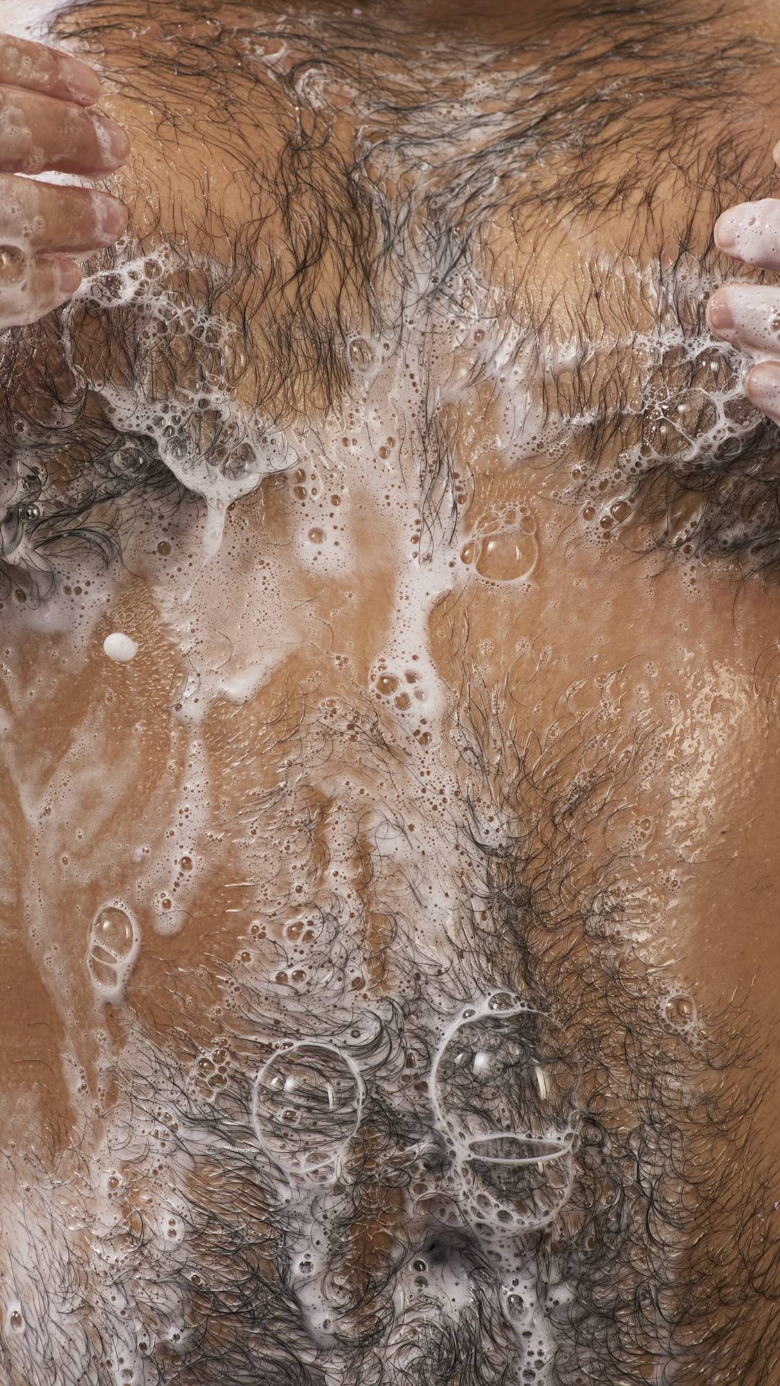 Close-up of soapy lather with delicate white bubbles spread across a person's wet, hairy chest during a shower.