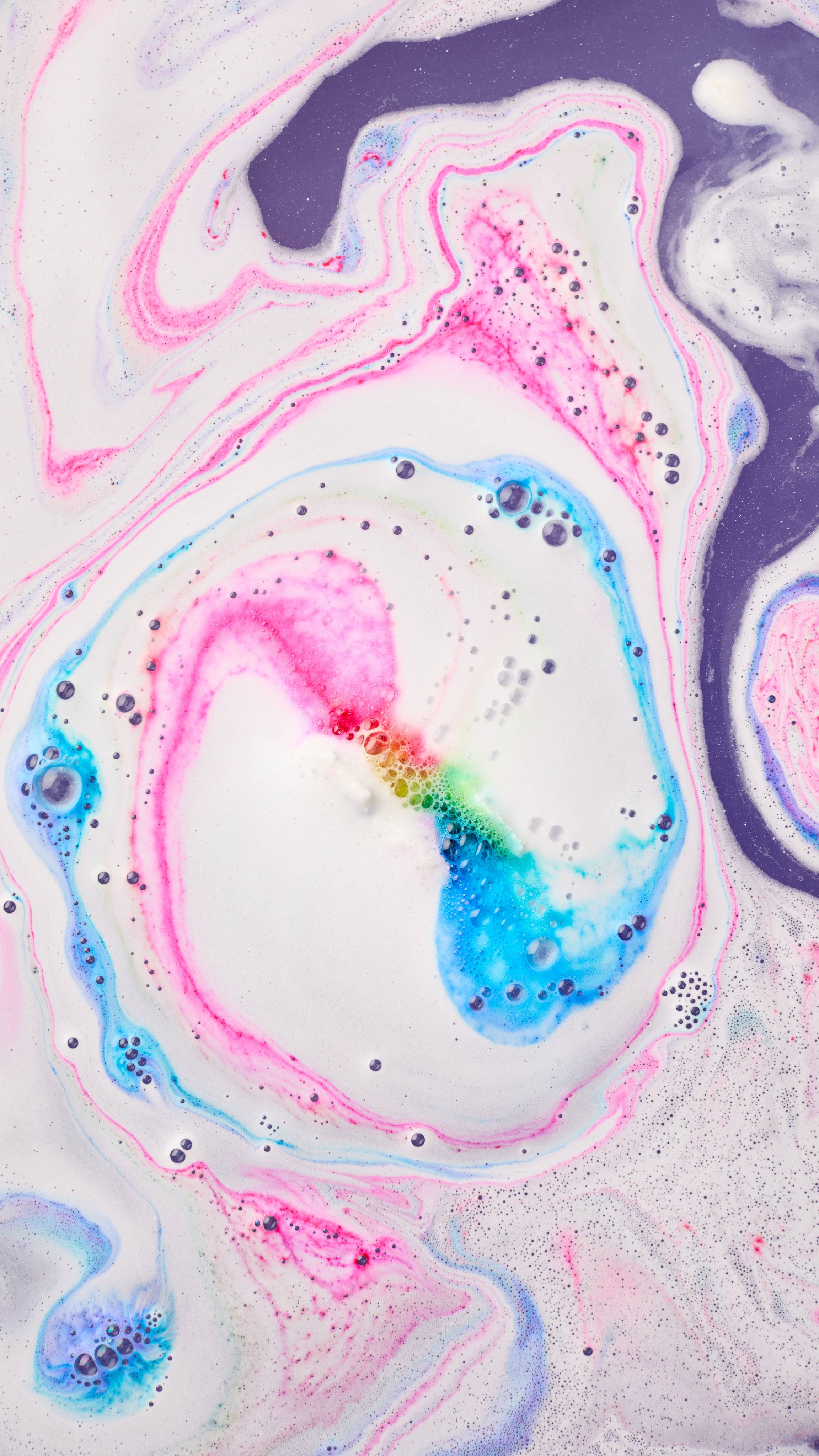 A close-up view of swirling bath foam in vibrant bright pink, cyan blue, green, and yellow, forming dynamic rainbow loops across milky white and deep purple water.