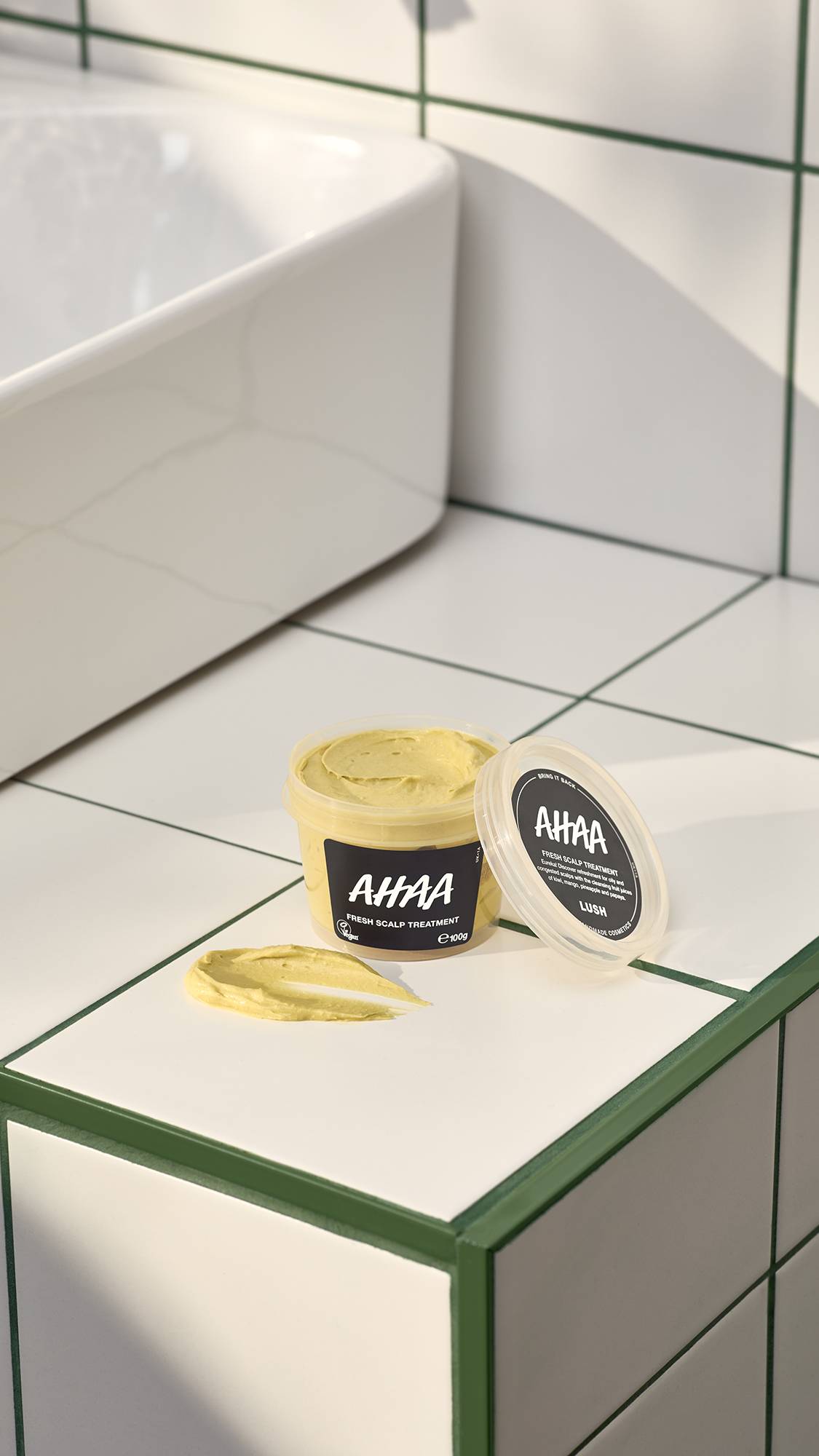 An open pot of Lush "Ahaa" fresh scalp treatment sits on a white tiled surface with green grout. Beside the pot and its lid is a thick smear of the mustard-yellow, grainy treatment. A white sink is partially visible in the background.