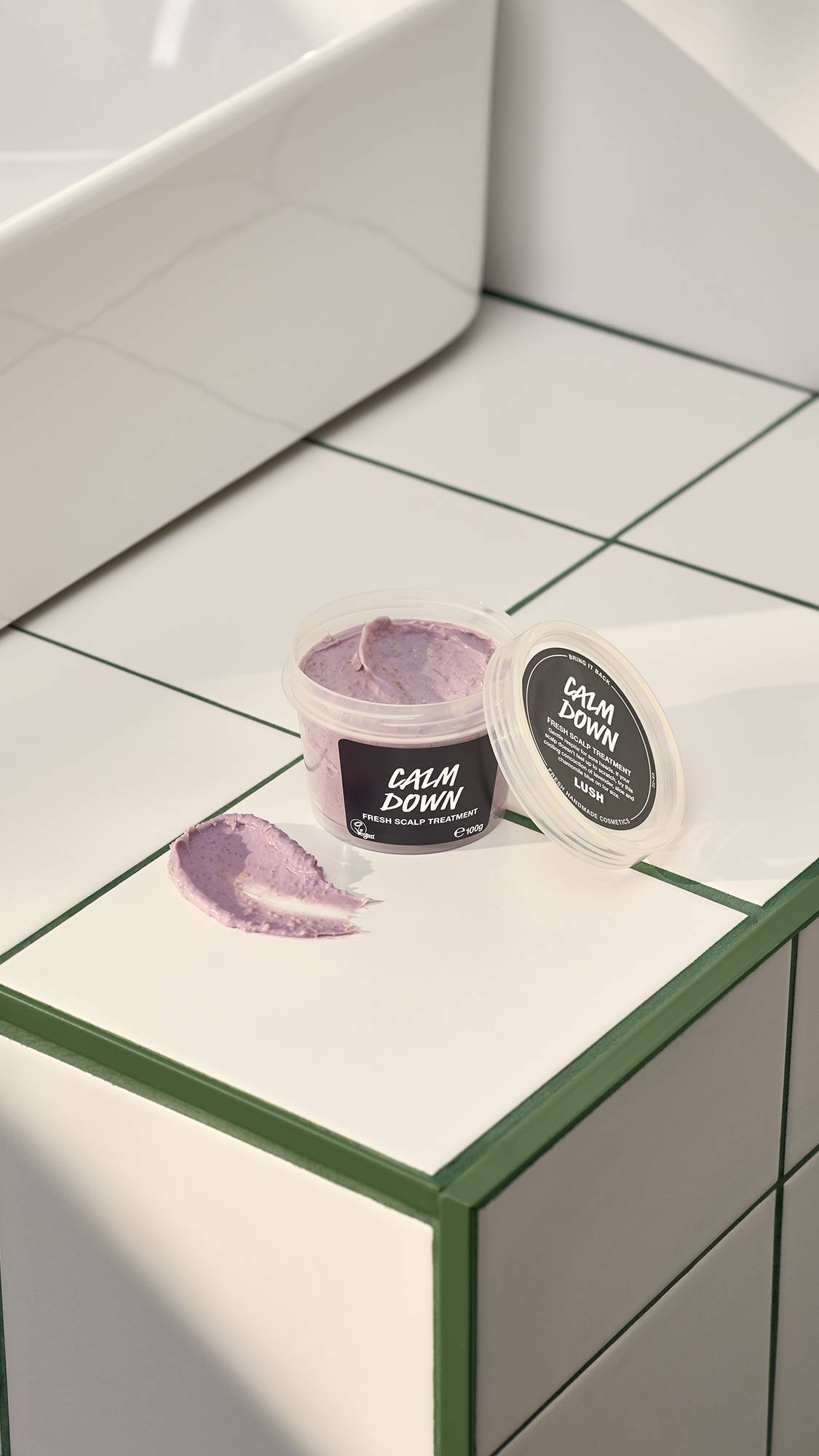 An open pot of Lush "Calm Down" fresh scalp treatment sits on a white tiled surface with green grout. Beside the pot and its lid is a thick smear of the grainy, lilac-coloured treatment. A white sink is partially visible in the background.
