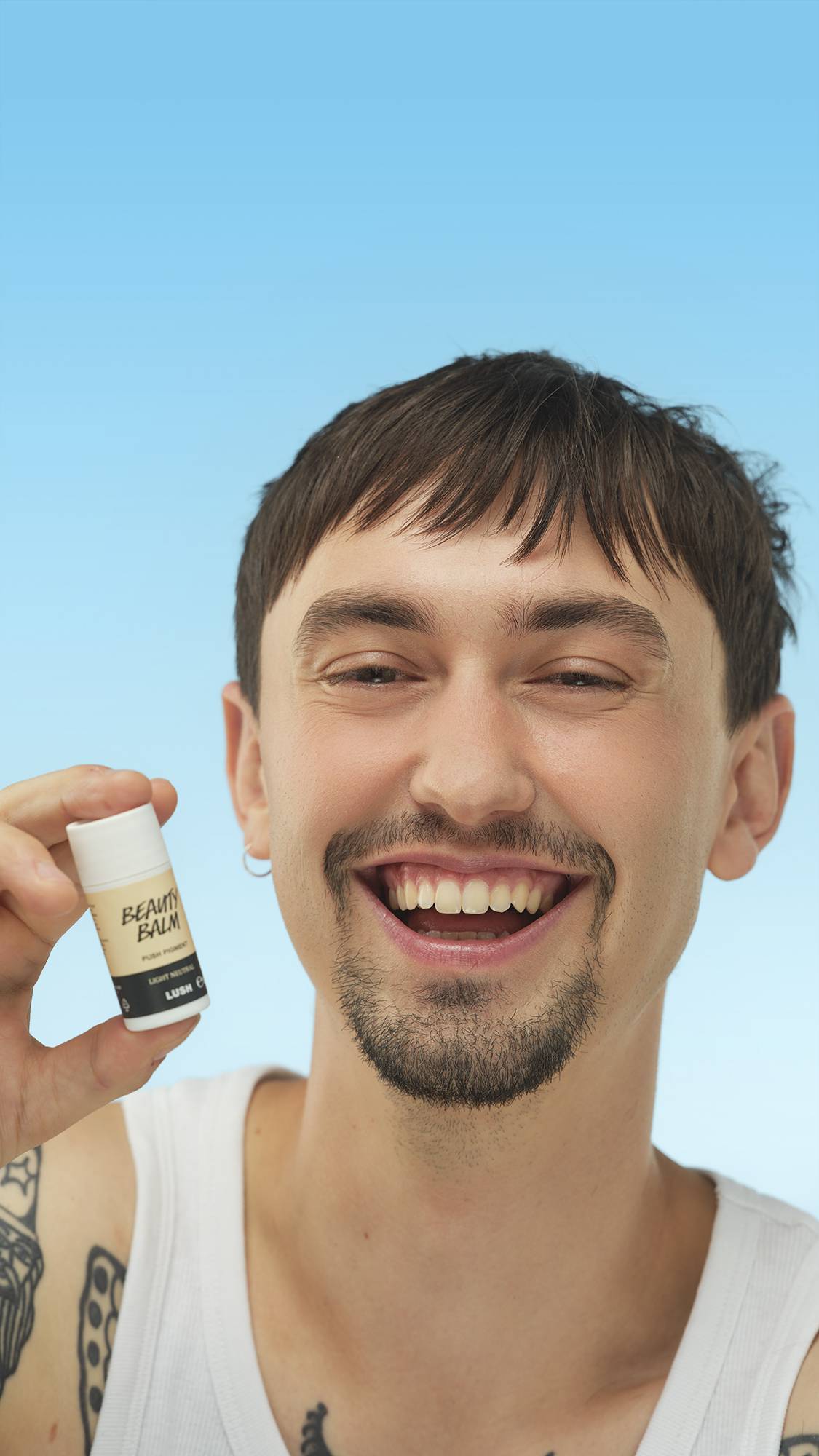 A close-up vertical portrait of a broadly smiling person with light-medium skin, holding the white tube of Light Neutral Beauty Balm near their cheek. They have dark hair, a goatee, and an earring, set against a bright blue background.