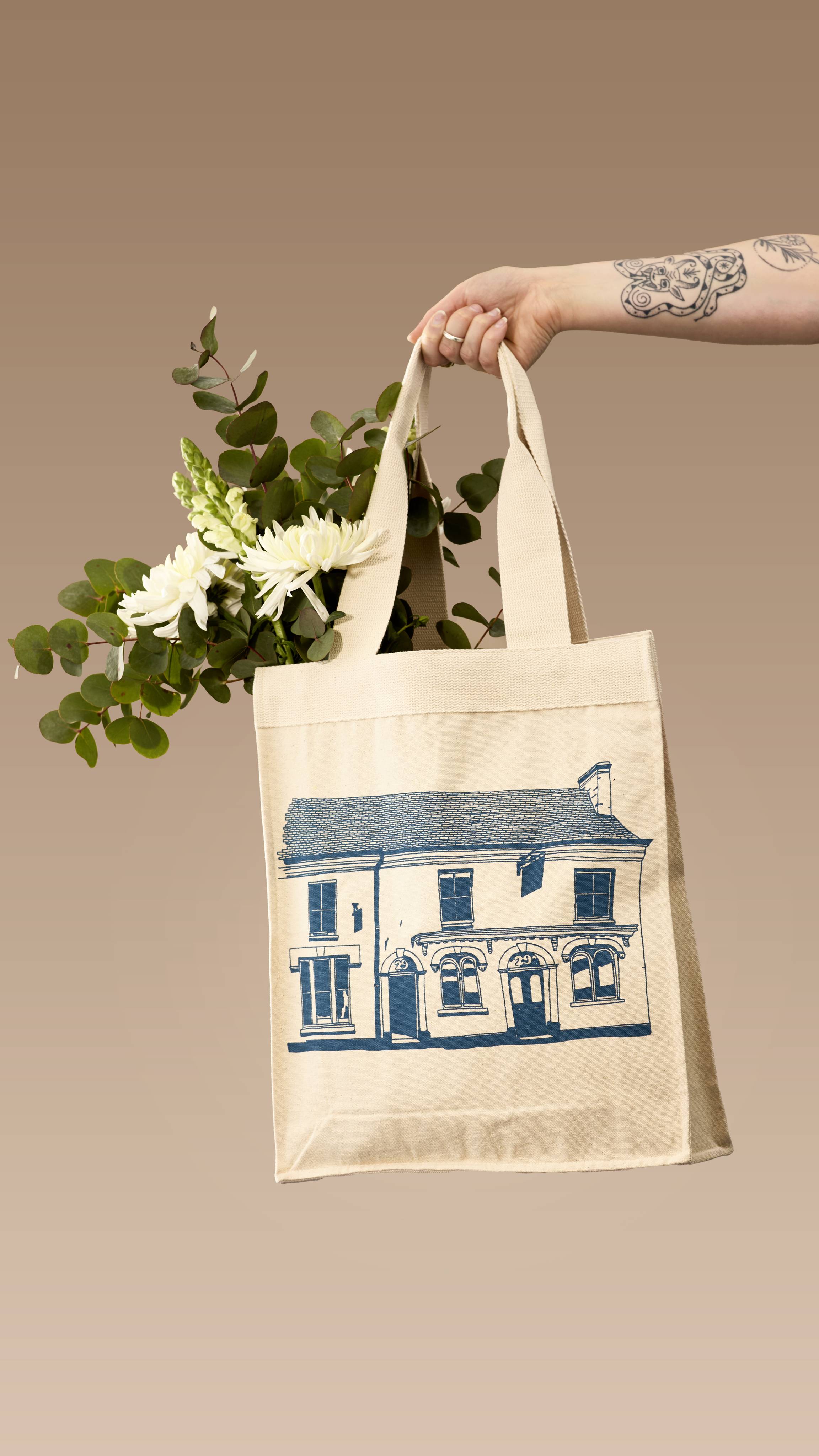 A tattooed arm holds up a cream canvas tote bag filled with white flowers and eucalyptus. The bag features a navy-blue line drawing of the original 29 High Street Lush shop facade, set against a neutral beige background.
