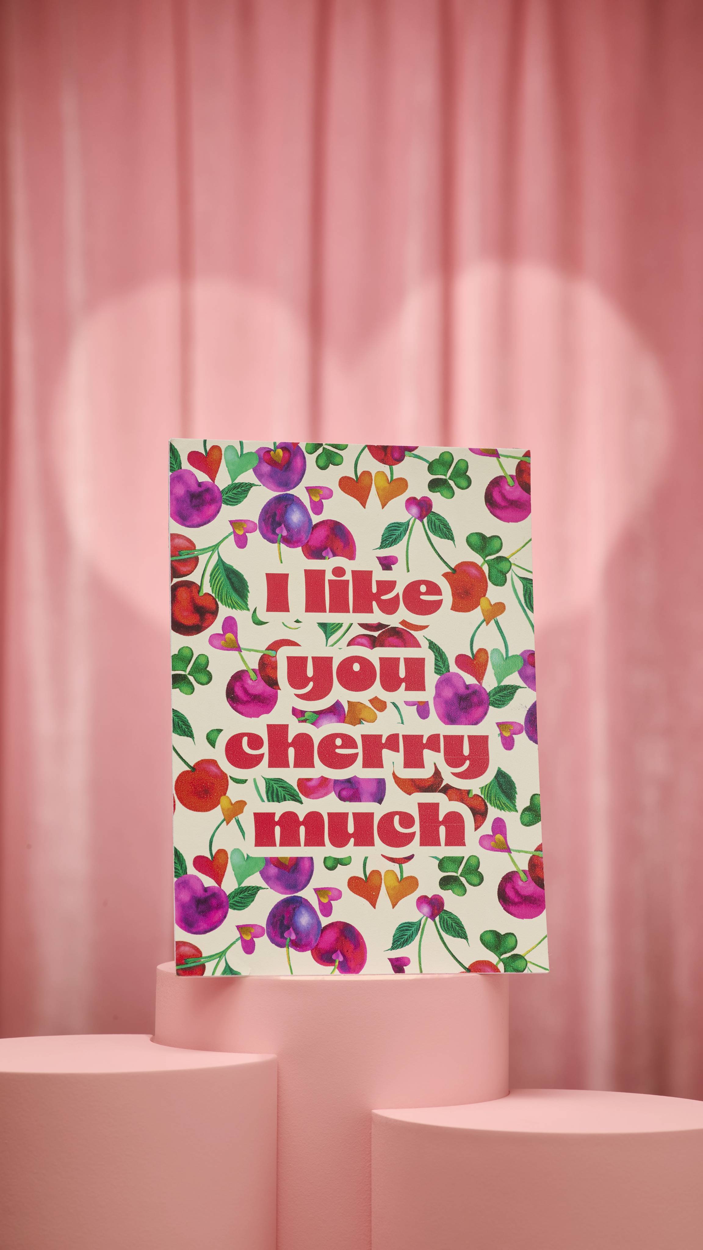 The "I Like You Cherry Much" card stands on a pink pedestal, displaying a cream background with a pattern of heart-shaped cherries in red, pink, and purple. Bold, glittery red text reads "I like you cherry much".