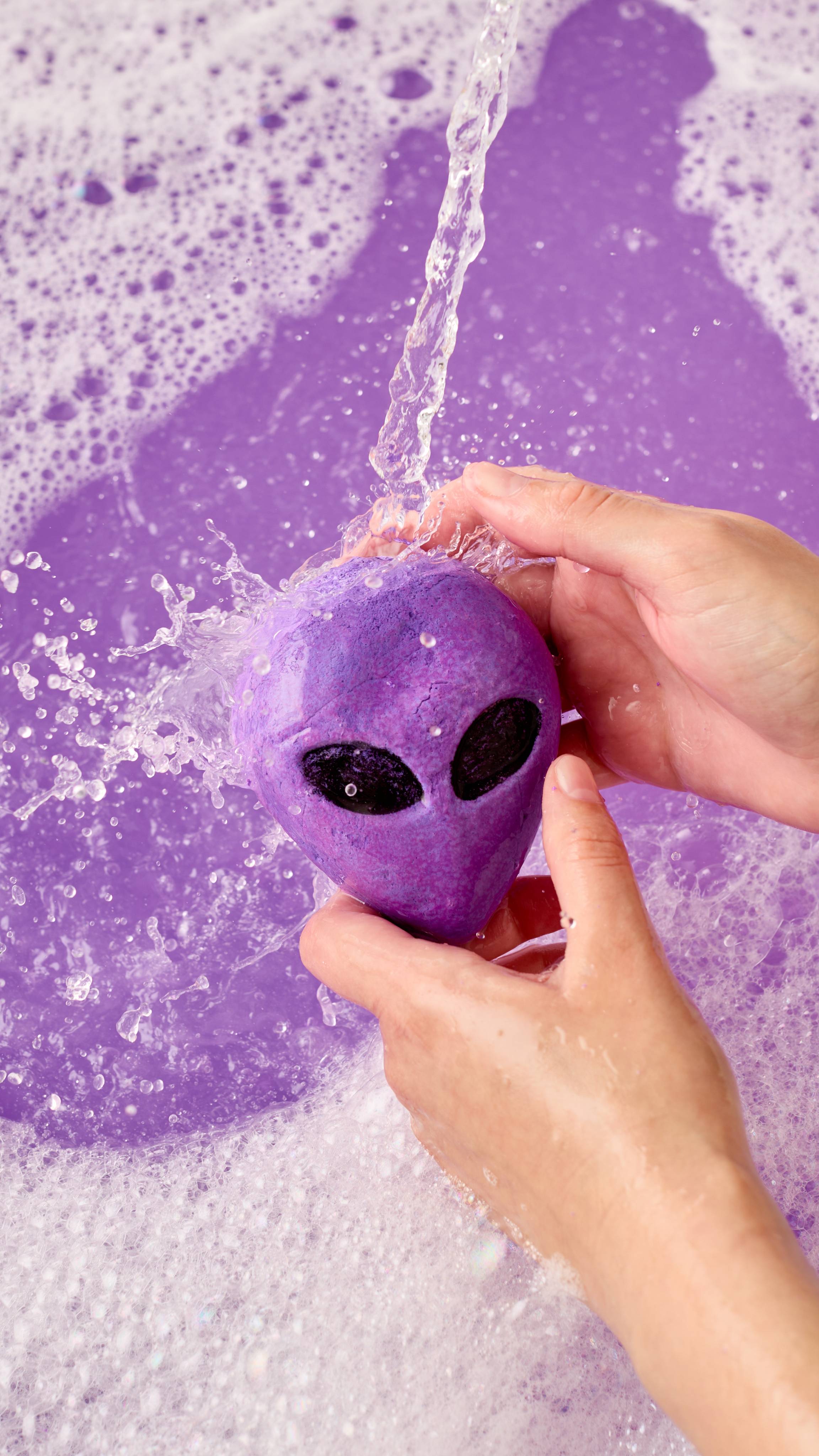 The image shows two light-toned hands holding a vibrant purple 'Alien' face-shaped bubble bar under running water. A stream pours onto the bar, generating abundant thick foam across purple bath water.