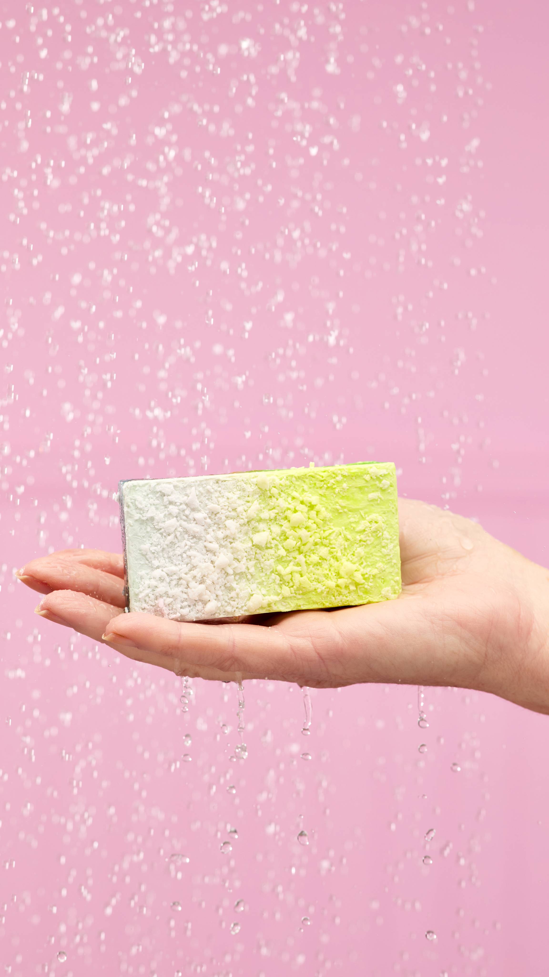 A hand holds a rectangular Baked Alaska soap slice under a stream of water in a shower, on a pale-pink background. The soap is on its side with the white, frosted top facing the camera. 