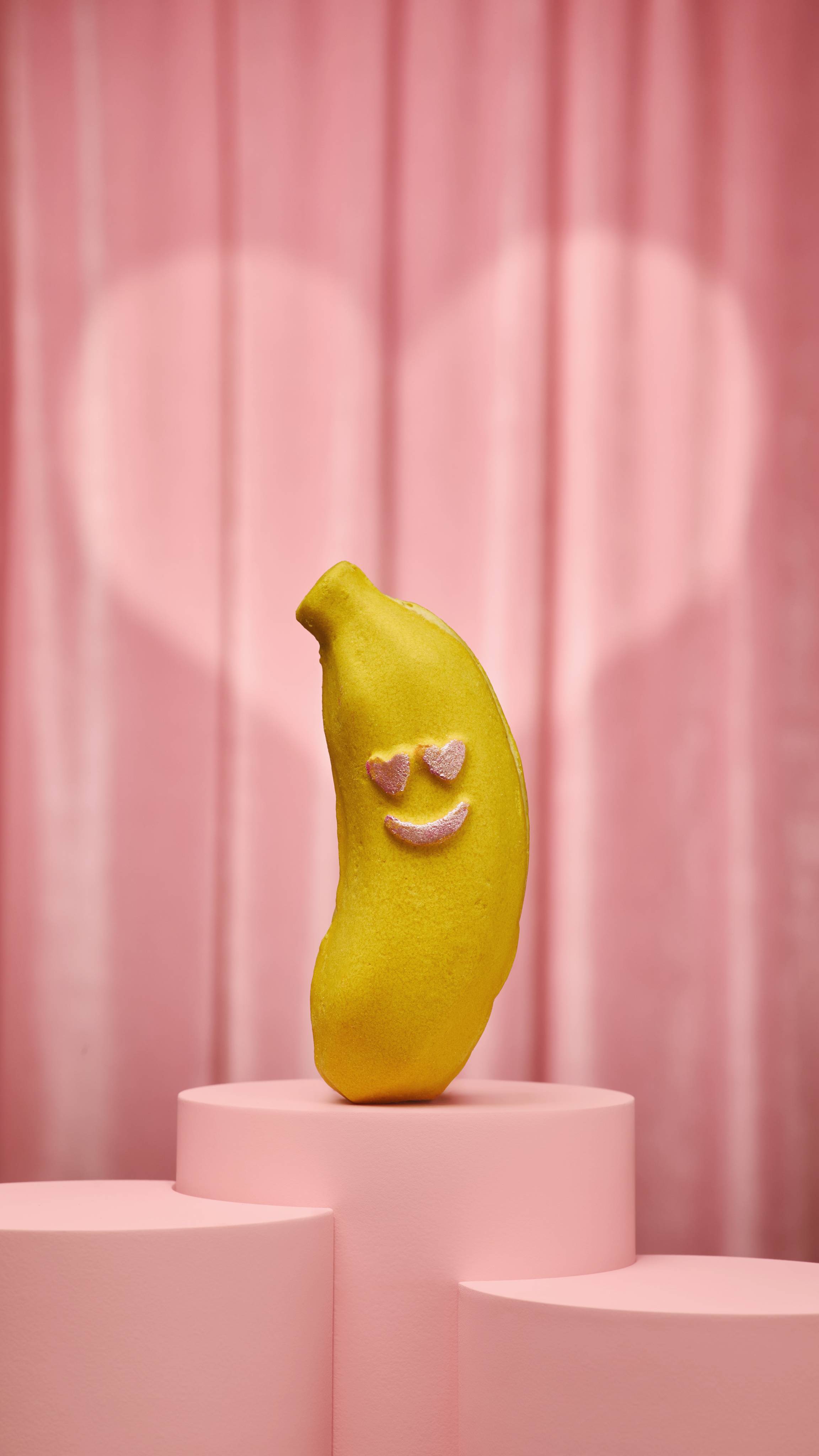 The image shows a yellow, banana-shaped bubble bar with a smiley face consisting of pink heart eyes and a curved mouth. The product sits on a pink podium on a pink fabric background.