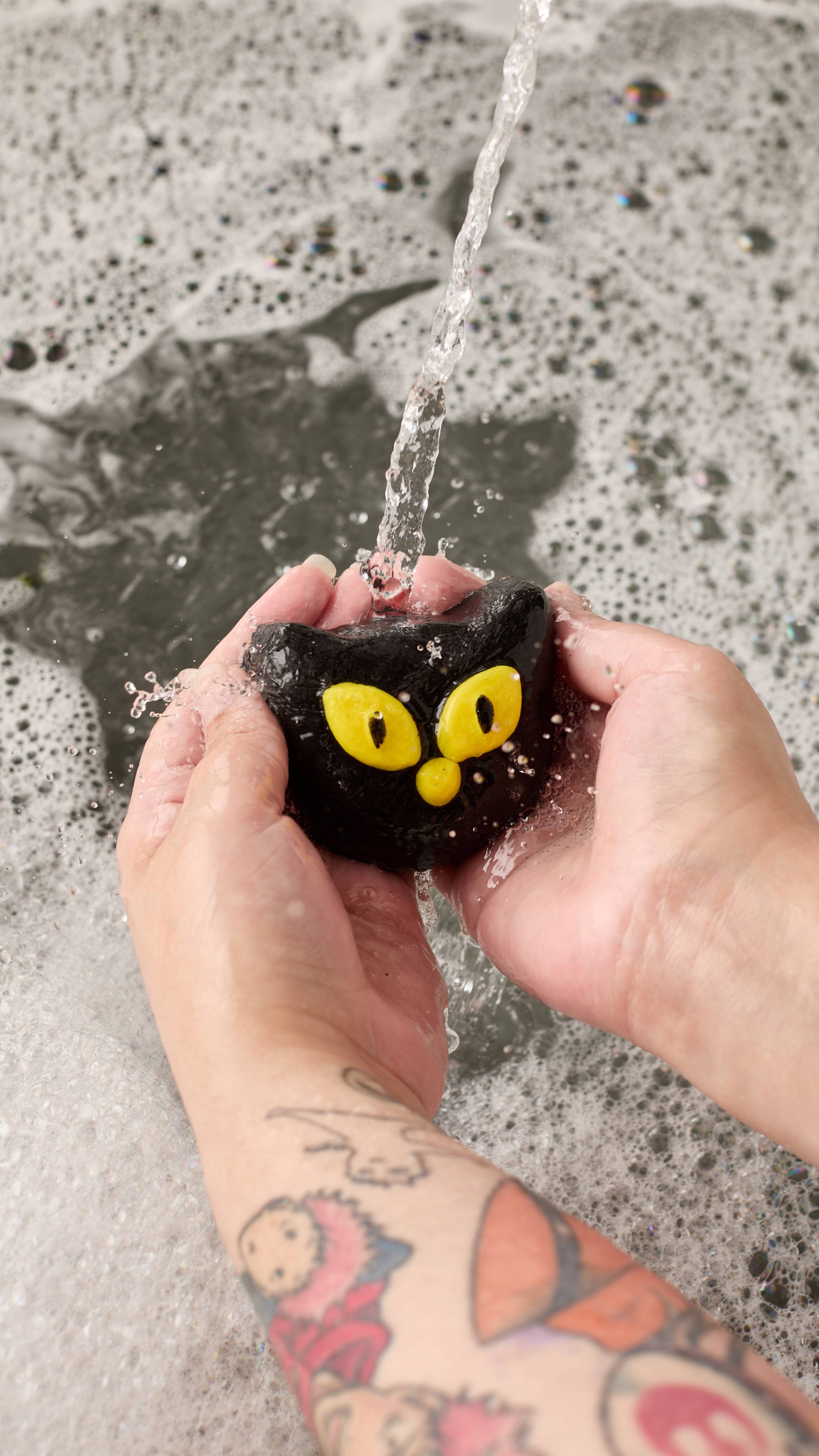 The image shows two light-toned hands holding the 'Bewitched' bubble bar in the shape of a black cat's face with yellow eyes and nose under running water. A stream pours onto the bar, generating thick bubbles on the dark bathwater.