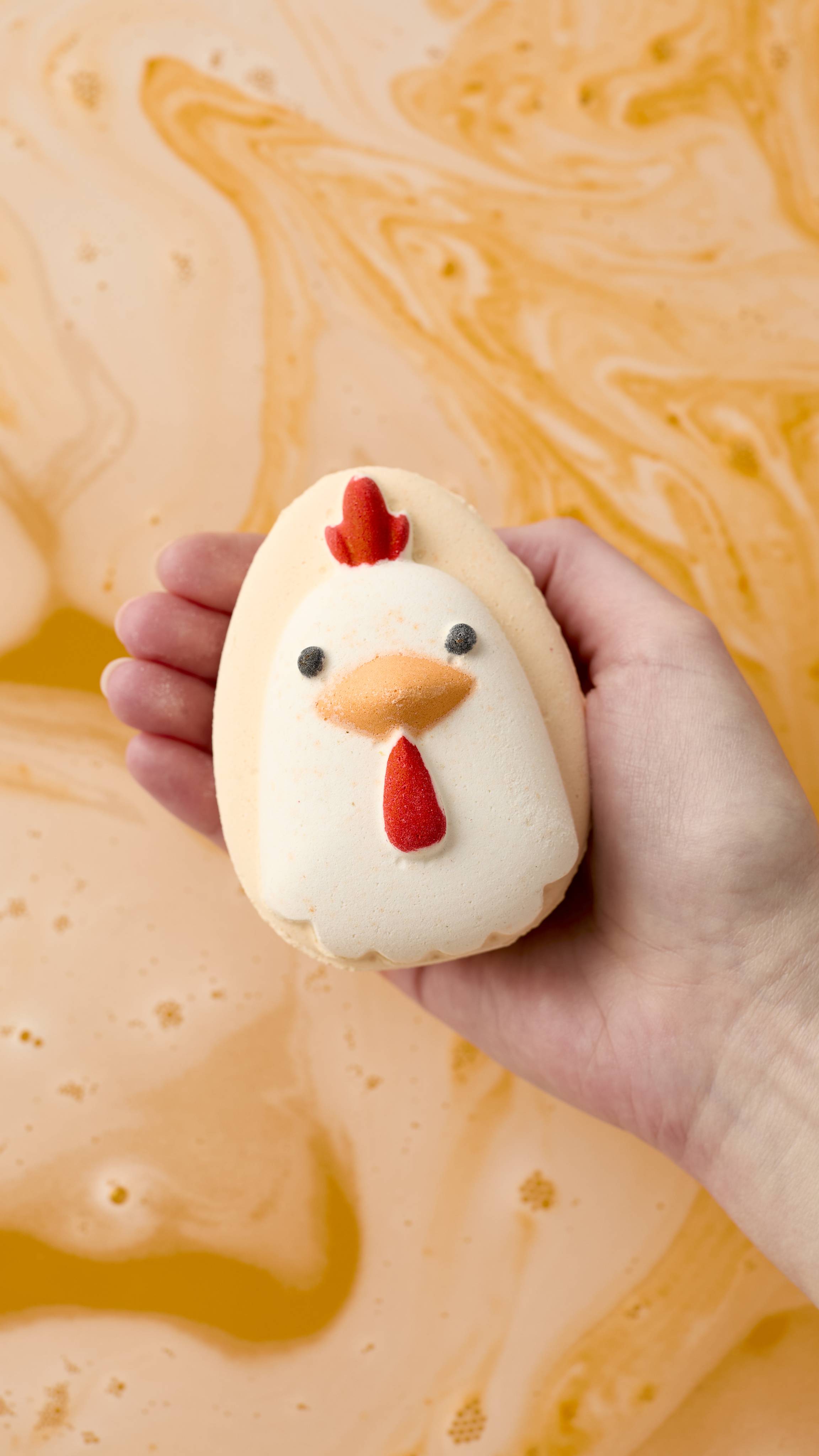 An aerial view image of a model's hand holding the "Big Dilemma" Lush Easter bath bomb above swirling orange bathwater. The egg-shaped bath bomb has the head of a cockerel on top, complete black eyes, an orange beak and red details.