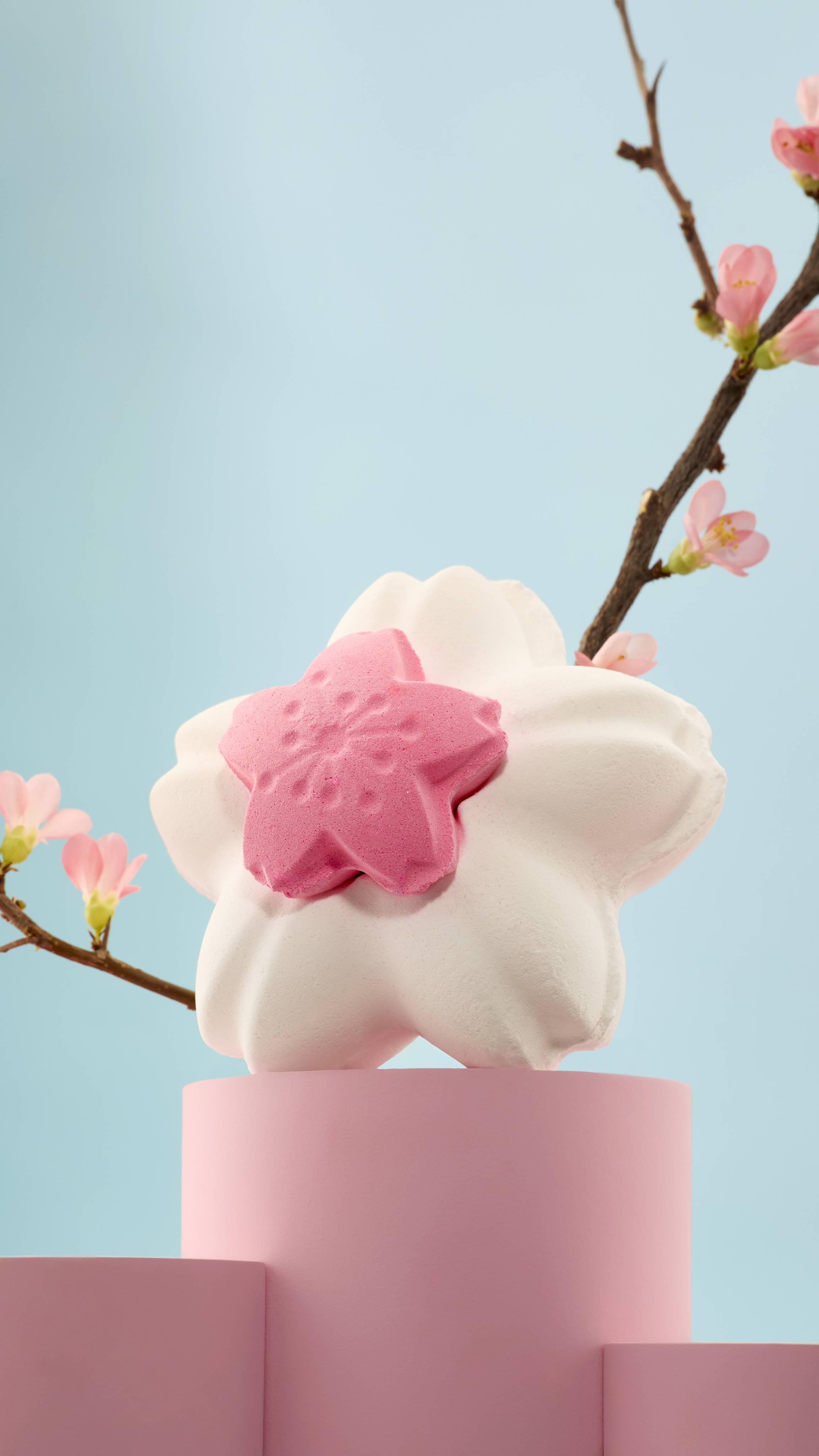 A Blooming Cherry Blossoms bath bomb, with a small pink flower centred on a larger white one, sits on a pink podium. A real blossoming branch curves behind it against a soft blue background. 