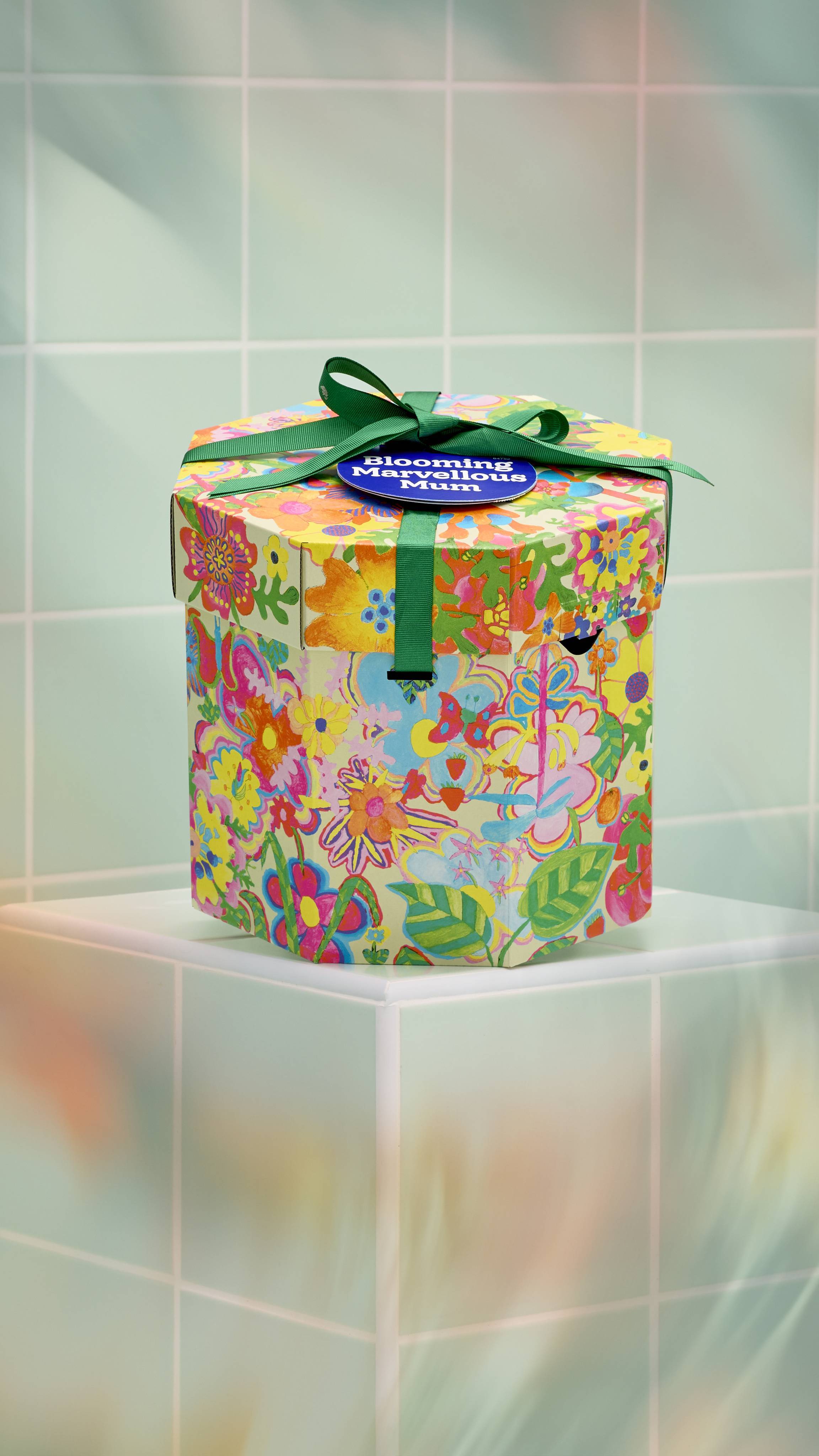 A vibrant, hexagonal gift box with a colourful, hand-drawn floral pattern sits on a pale green tiled surface. It is tied with a green ribbon and features a blue circular tag reading ‘Blooming Marvellous Mum’. 