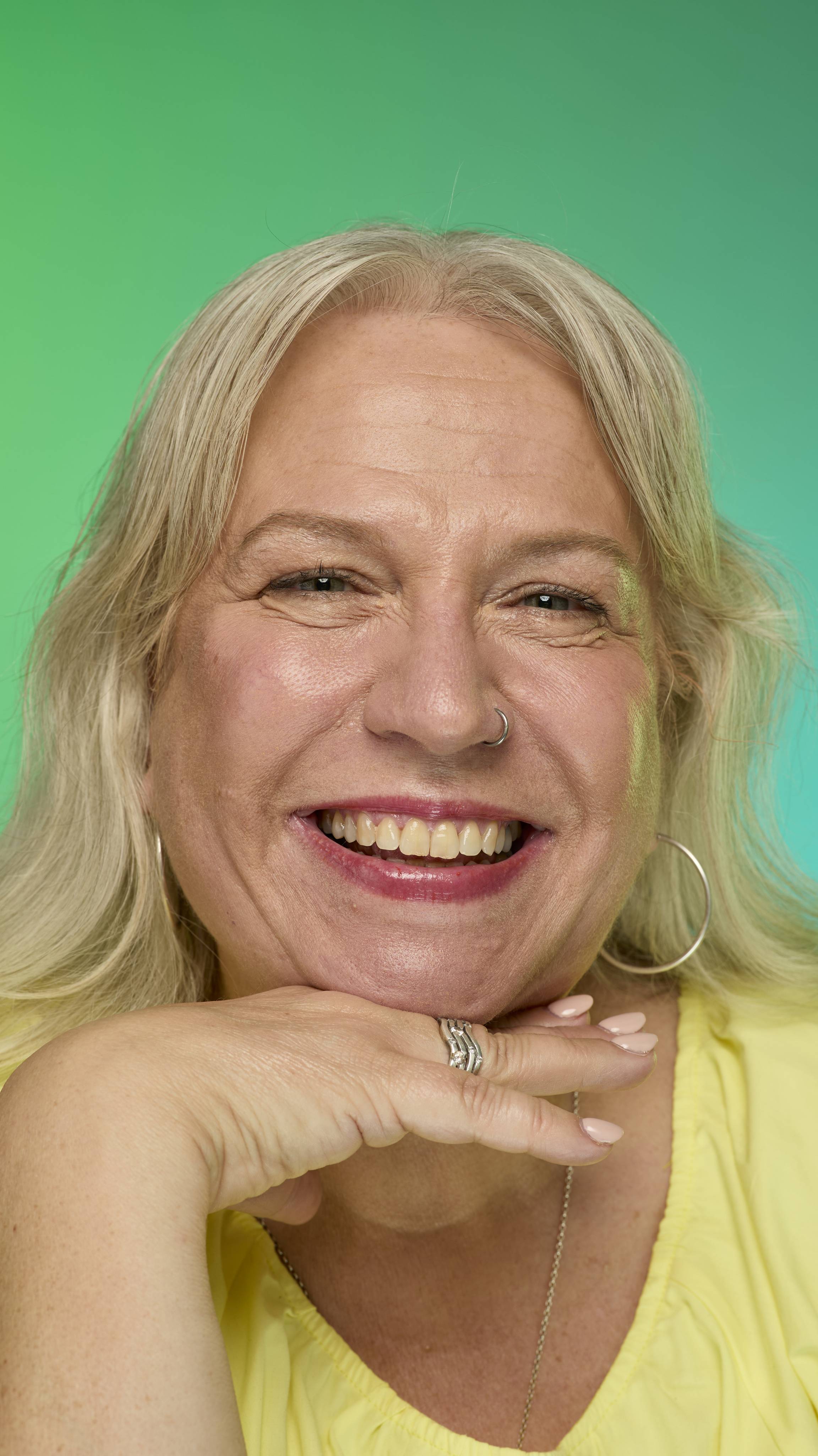 The image shows a model with blonde hair on a green and blue gradient background. They are smiling at the camera as their eyebrows look thick and full thanks to the Blonde brow pomade.