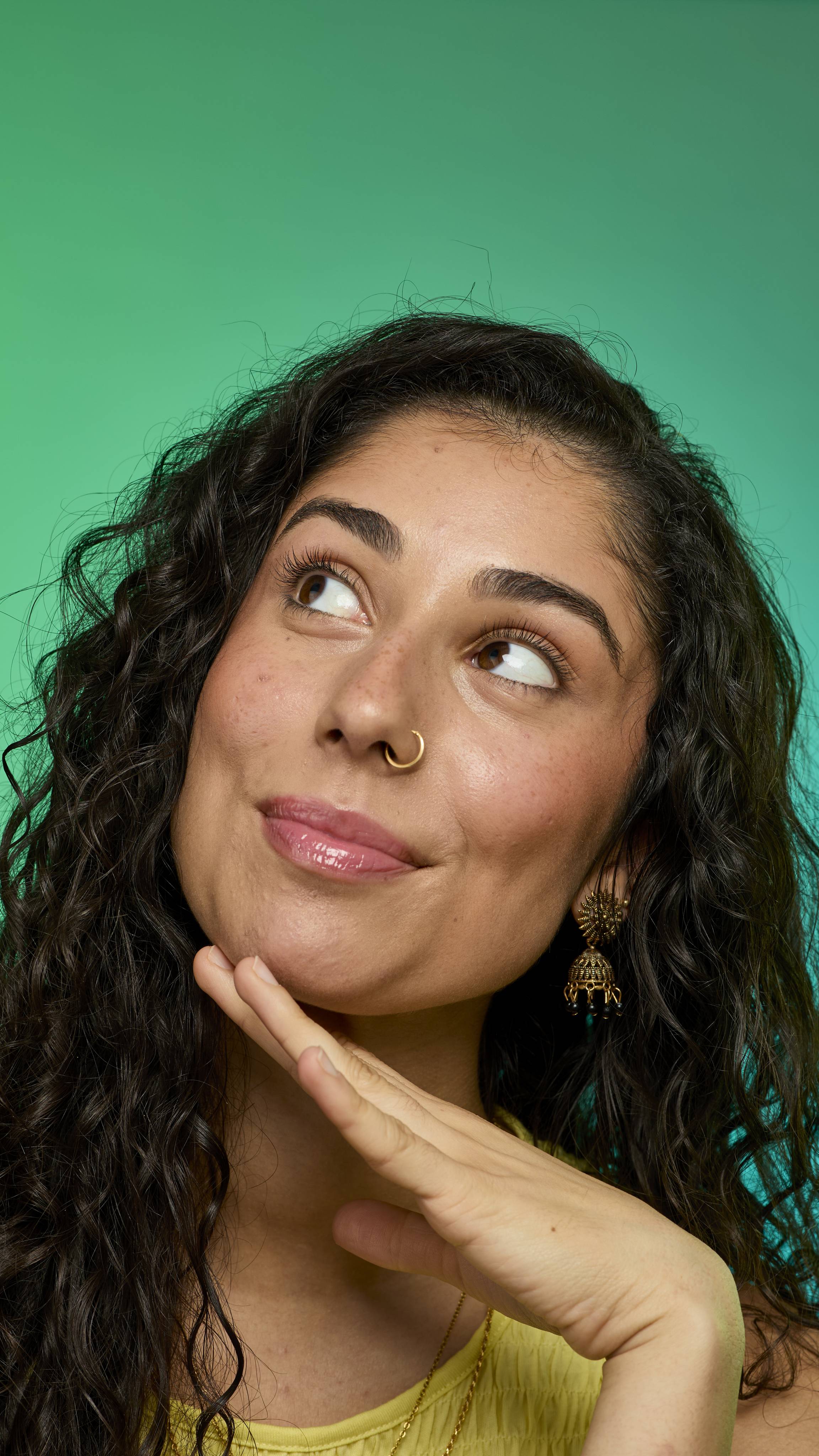 The image shows a model with dark curly hair on a green background. They are holding their hand up to their face as they glance away. Their eyebrows look thick and full thanks to the Brun brow pomade.