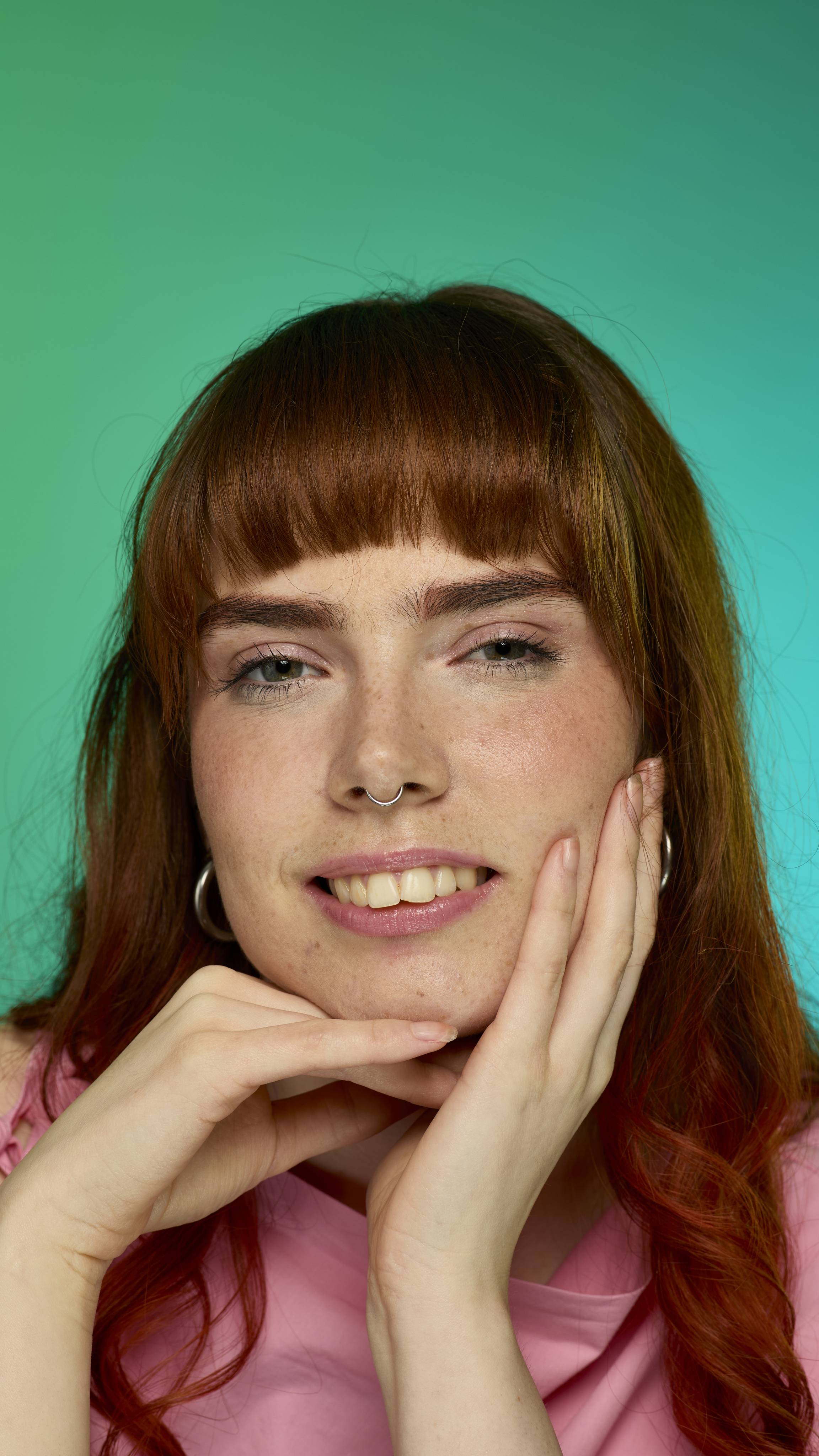 The image shows a model with auburn straight hair with a full fringe on a green and blue gradient background. They are smiling at the camera as their eyebrows look thick and full thanks to the Rouge brow pomade.