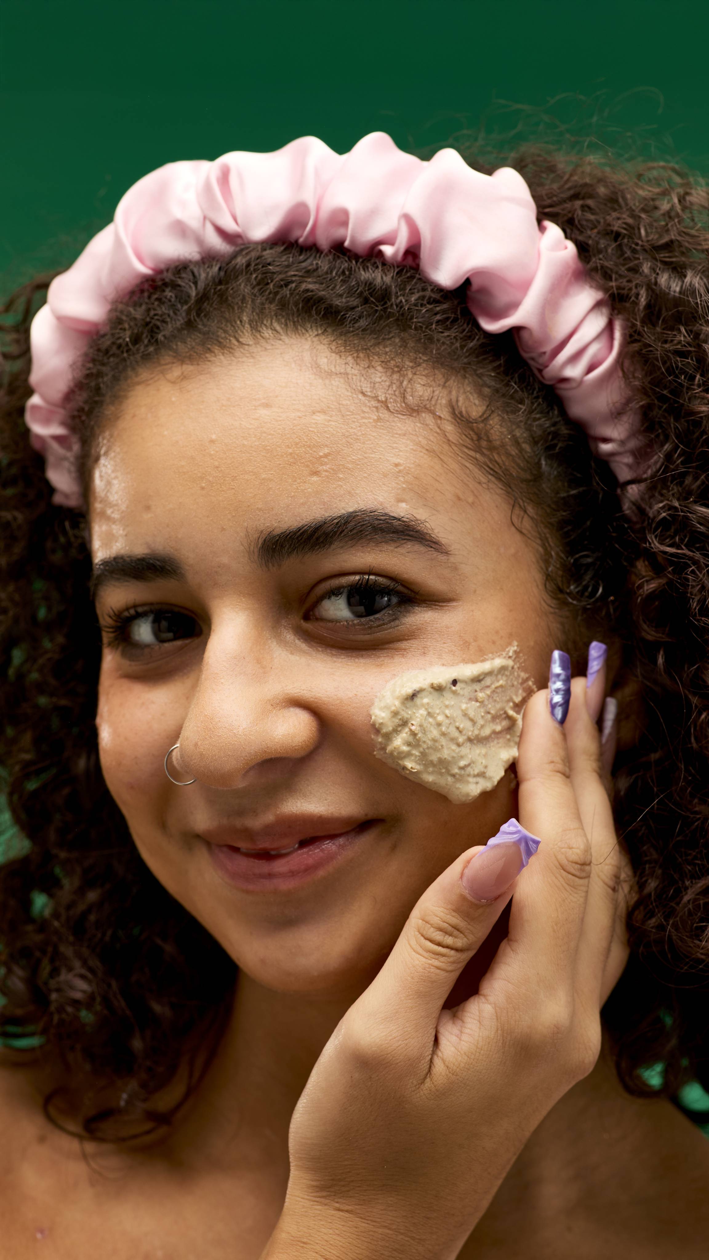 A person wearing a pink satin headband smiles while applying a LUSH Bûche de Noël cleanser to their cheek. The light-coloured, chunky cleanser is visible on their skin, and the background is a vibrant green.