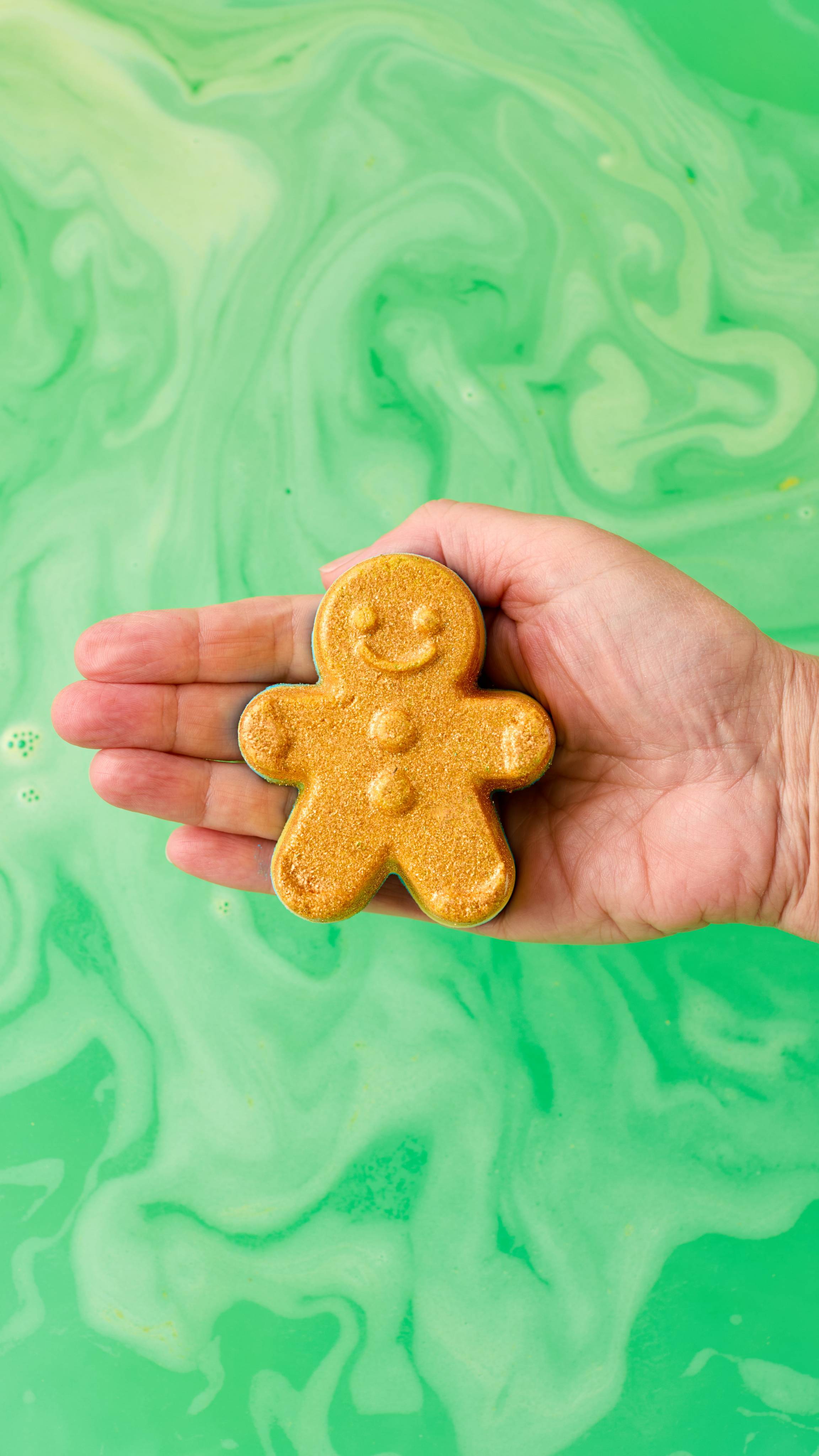 The image shows a light-toned hand holding a golden, Catch Me If You Can gingerbread man-shaped bath bomb with a smiling face. The product is held just above swirling green bath water.