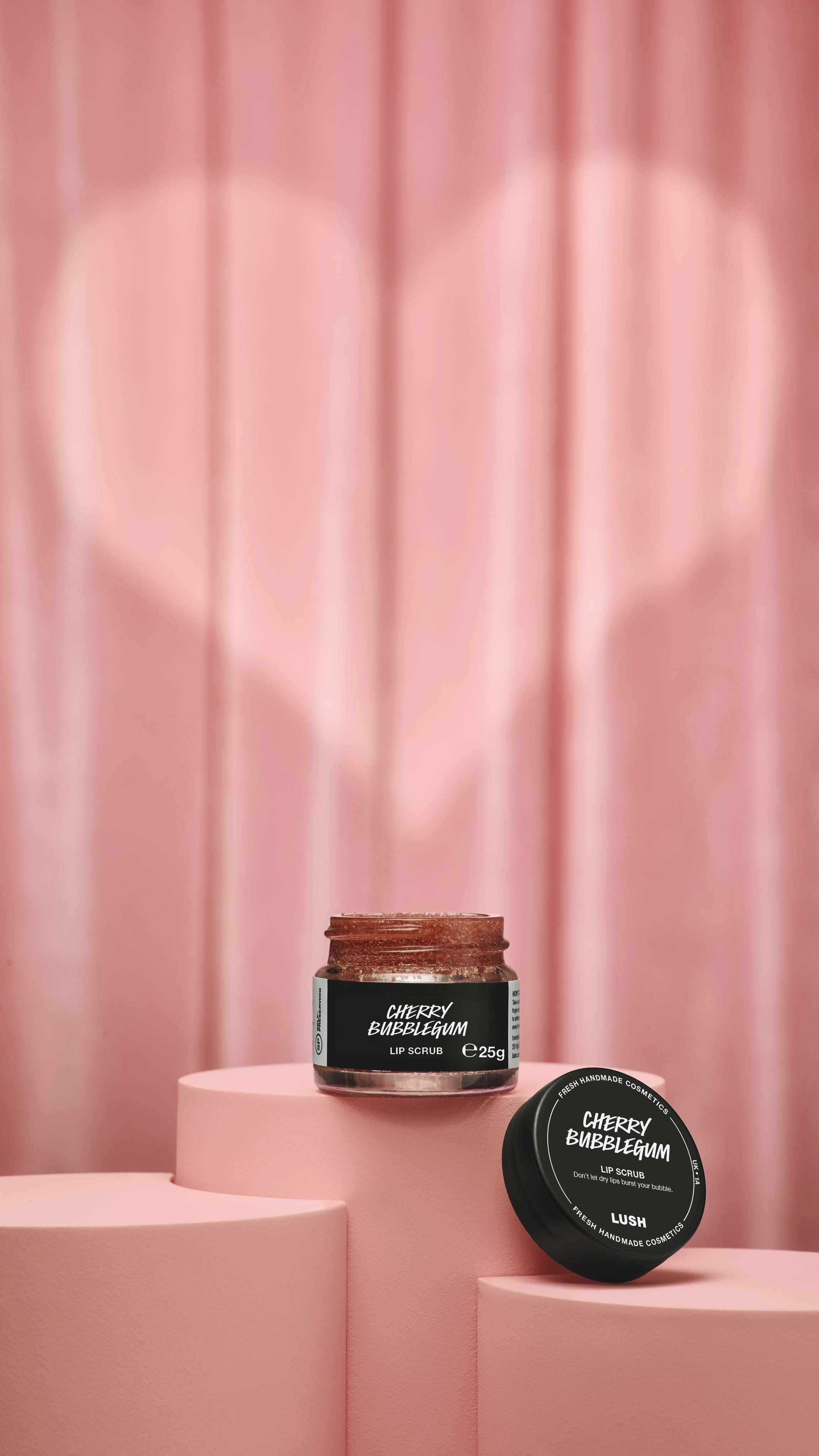 A small, clear glass jar of Lush "Cherry Bubblegum" lip scrub sits on a pink podium. The reddish-brown scrub is shown with its black lid resting nearby against a pink curtain with a heart-shaped spotlight.