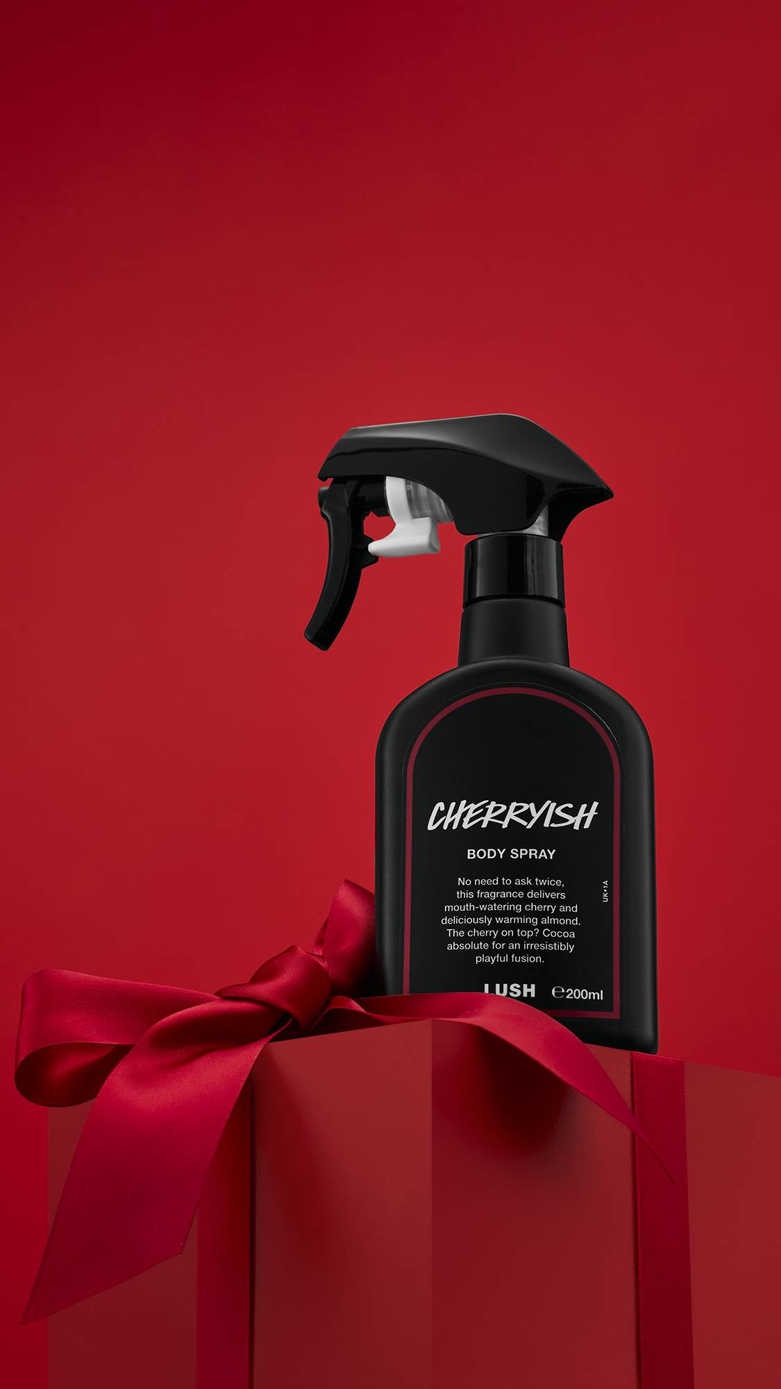 The image shows a solid red background with a matching red gift box tied with ribbon. The contracting black Cherryish body spray bottle sits on top of the gift. 