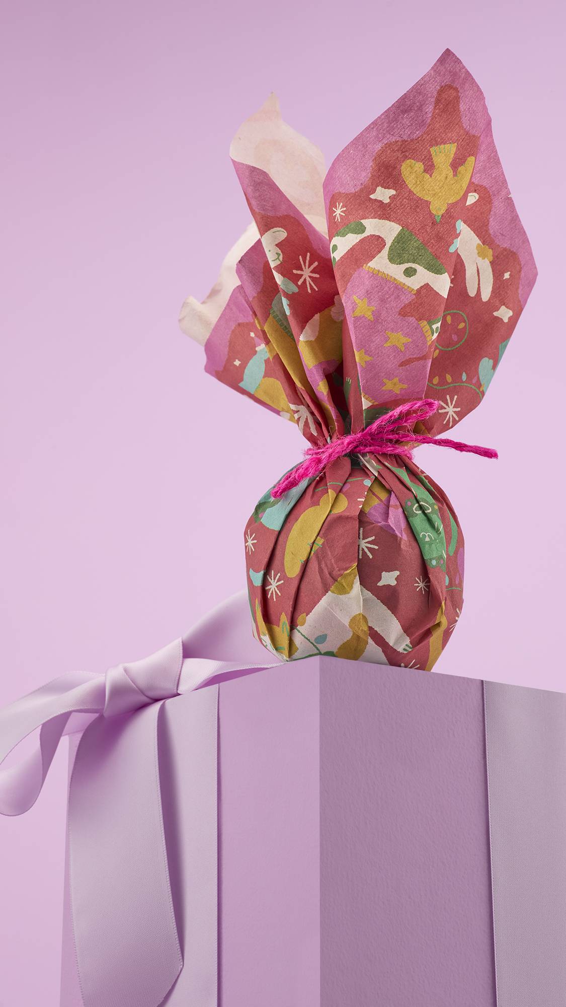 
A small, round gift wrapped in a colorful, patterned "Christmas Companions" lokta paper with reindeers and other festive characters. It's tied with pink string and sitting on a light purple, ribbon-wrapped box.