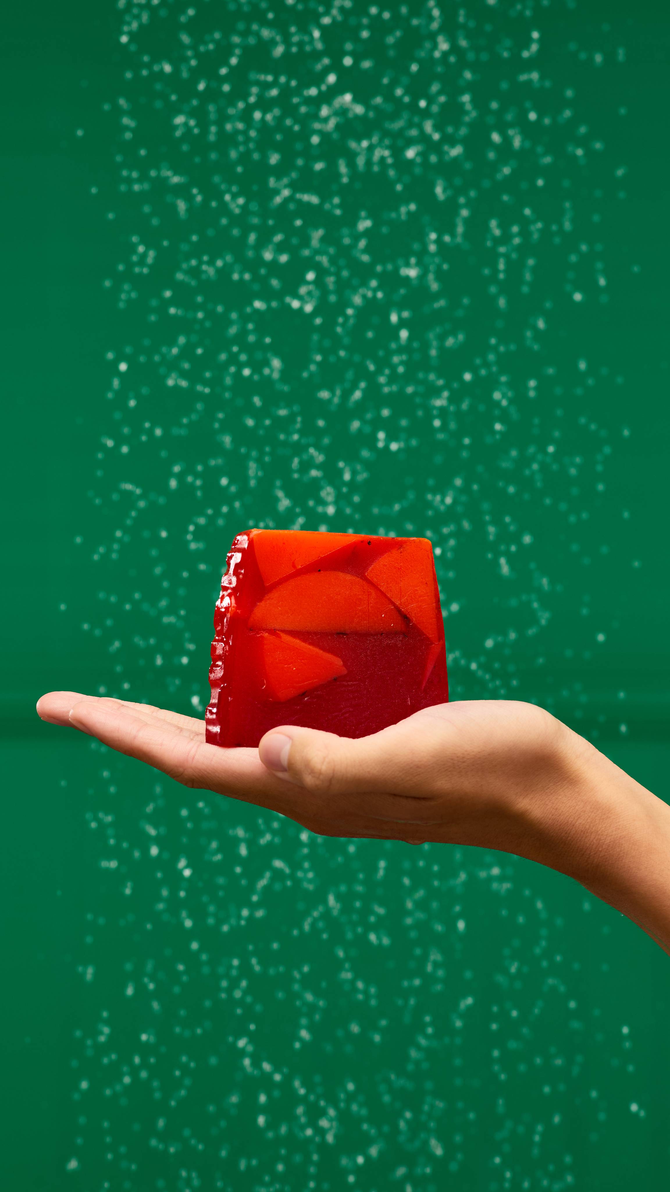 A fair-skinned hand holds a ran off-square slice of Christmas Cranberry soap. The red soap has solid lighter shapes throughout with dark flecks. It's held against a solid green background with a falling shower water behind.