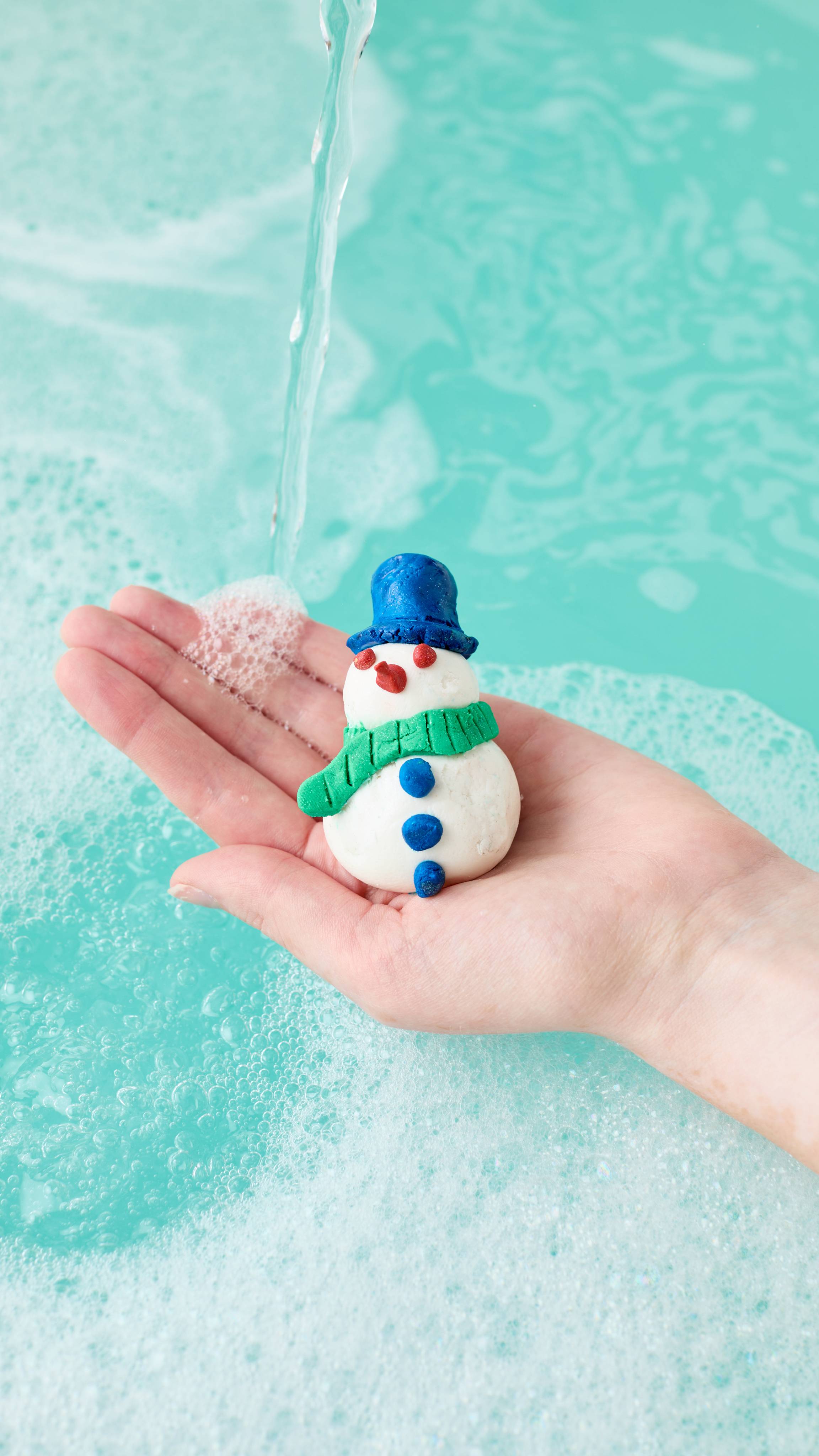 A hand holds the Christmas Lights Fun bath product, which has been moulded into the shape of a snowman, complete with blue hat, orange nose, green scarf and buttons. It is held under running water just above a frothy, teal-coloured bath.