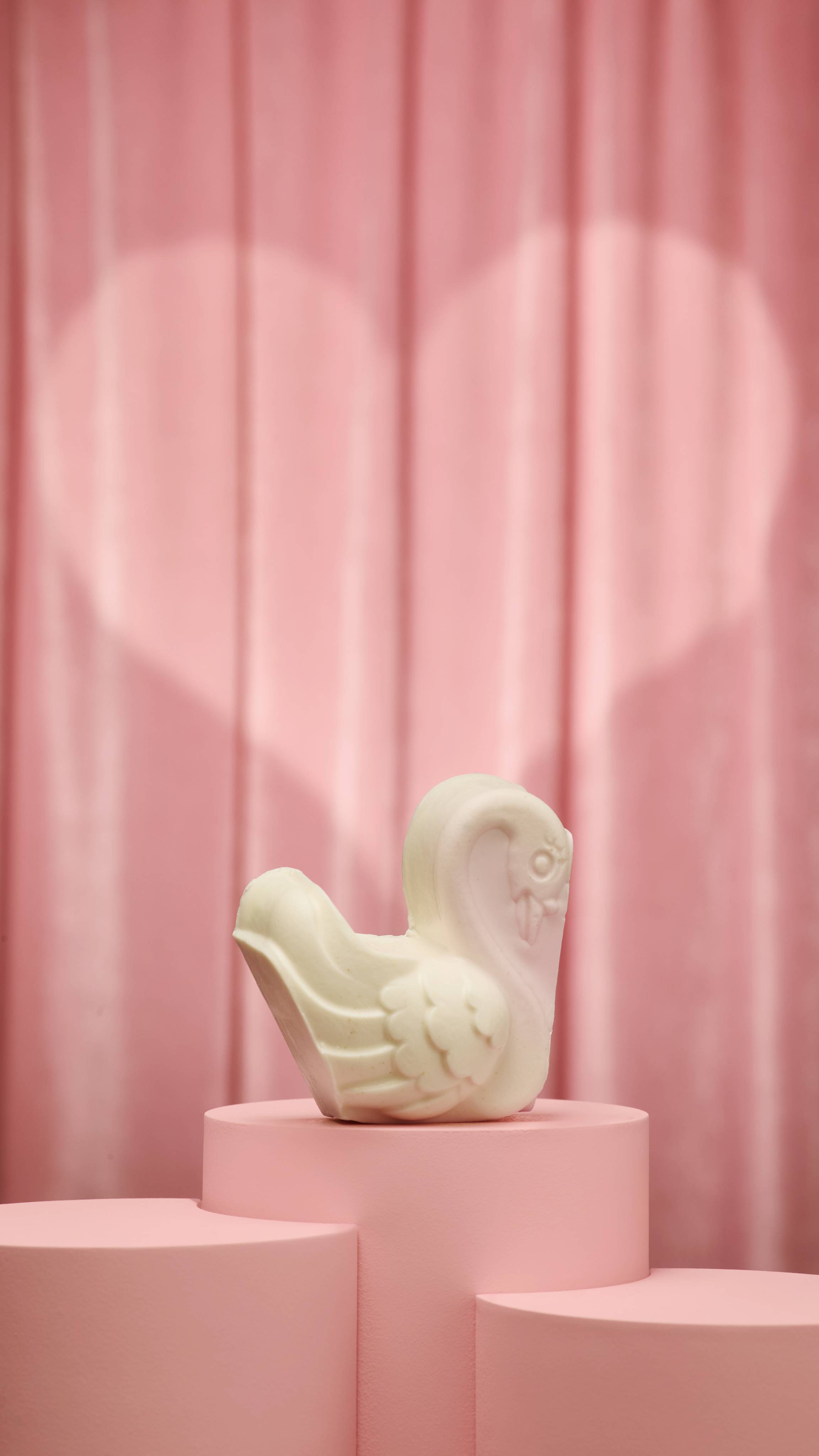 A white, single swan-shaped soap sits on a tiered pink podium against pink curtains with a heart-shaped spotlight. The swan features textured wing details and a subtle pink tint on its neck and head.