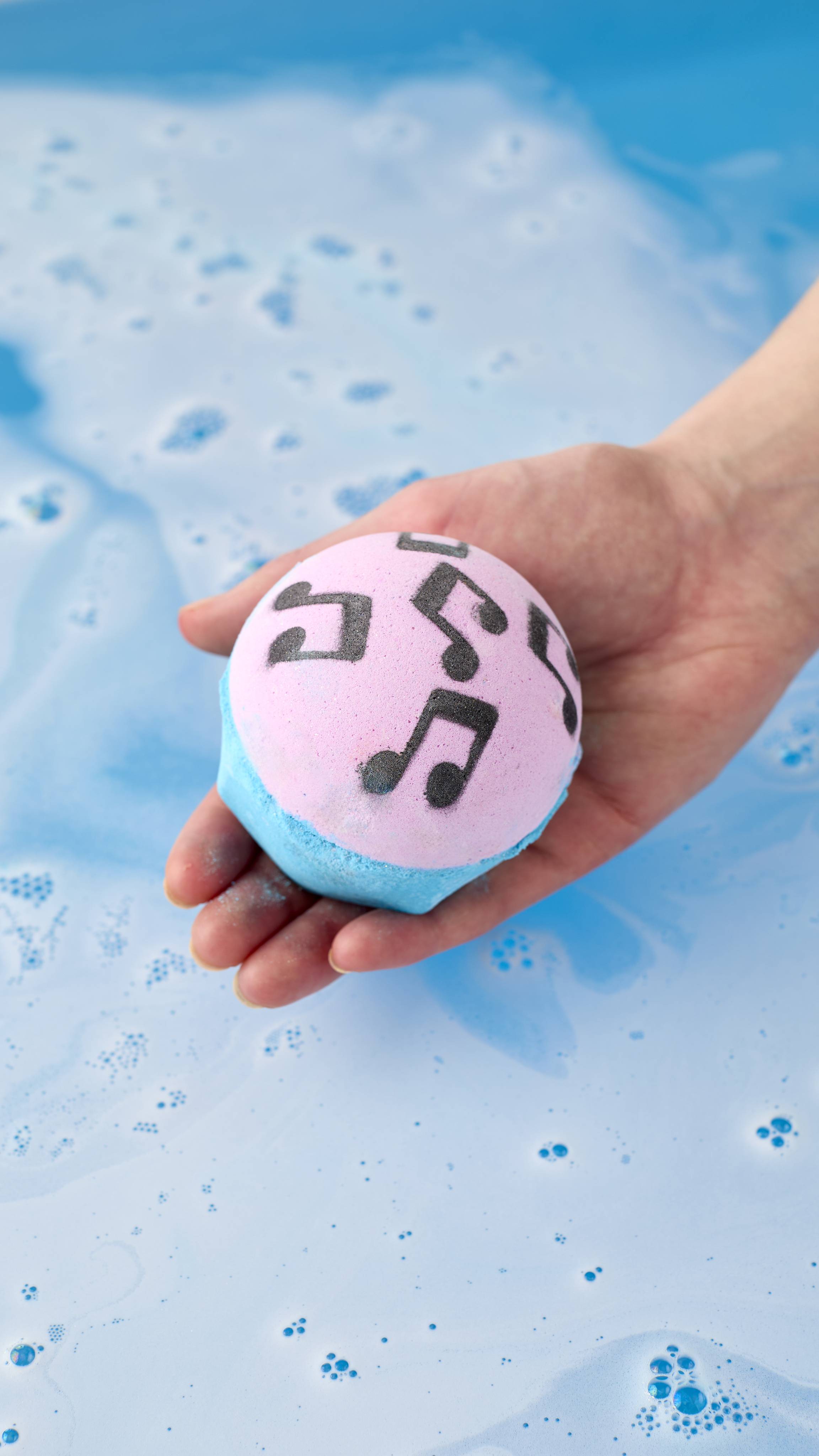 A model's open hand holds the pink and blue musical note "Dad Dancing" bath bomb over a bathtub. The water below is filled with pale blue and white soapy foam, with a few deeper blue bubbles scattered across the surface.