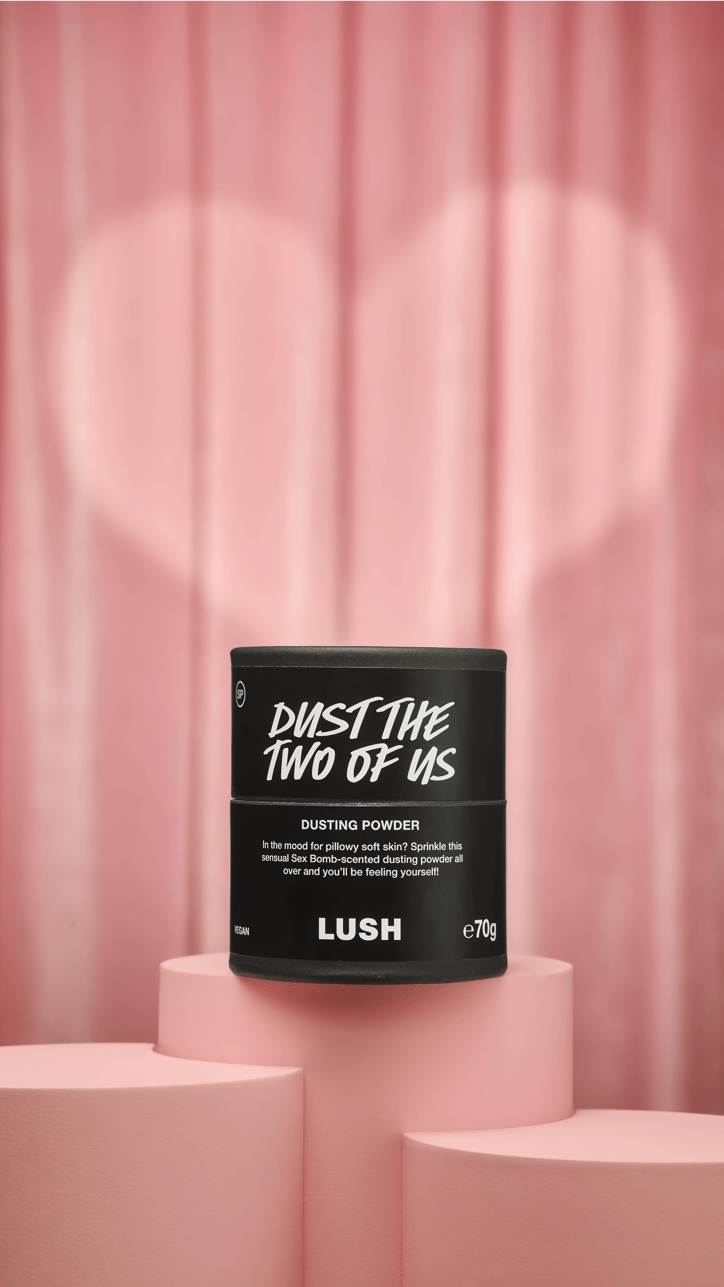 A black bottle of Dust The Two Of Us dusting powder rests on a pink pedestal against a soft pink curtain background illuminated by a heart-shaped spotlight.