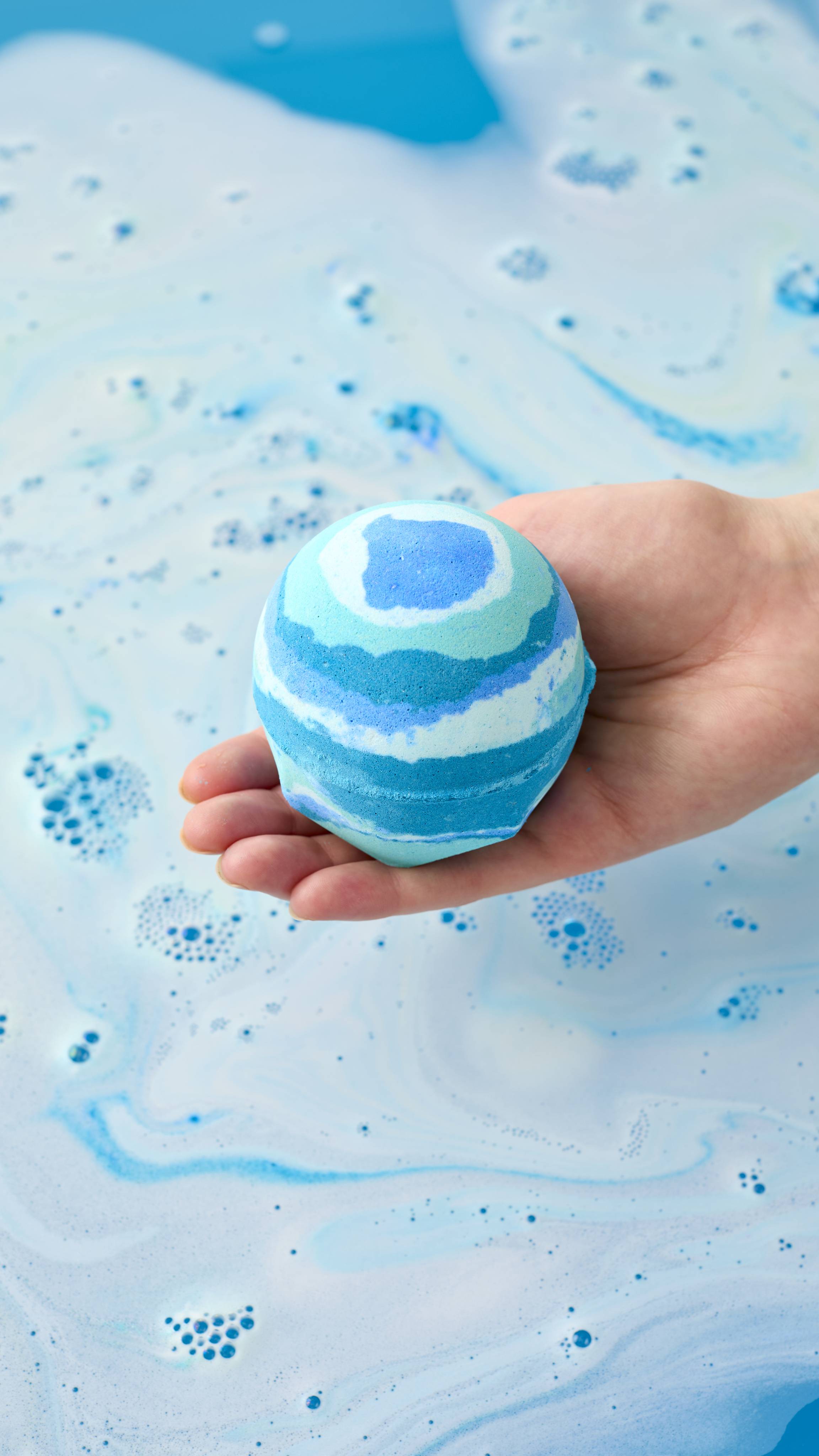 A model's open hand holds the Lush "Easy Breezy" bath bomb with blue and teal stripes over a bathtub. The water below is filled with pale blue and white foam, with swirling patterns and clusters of small bubbles on the surface.
