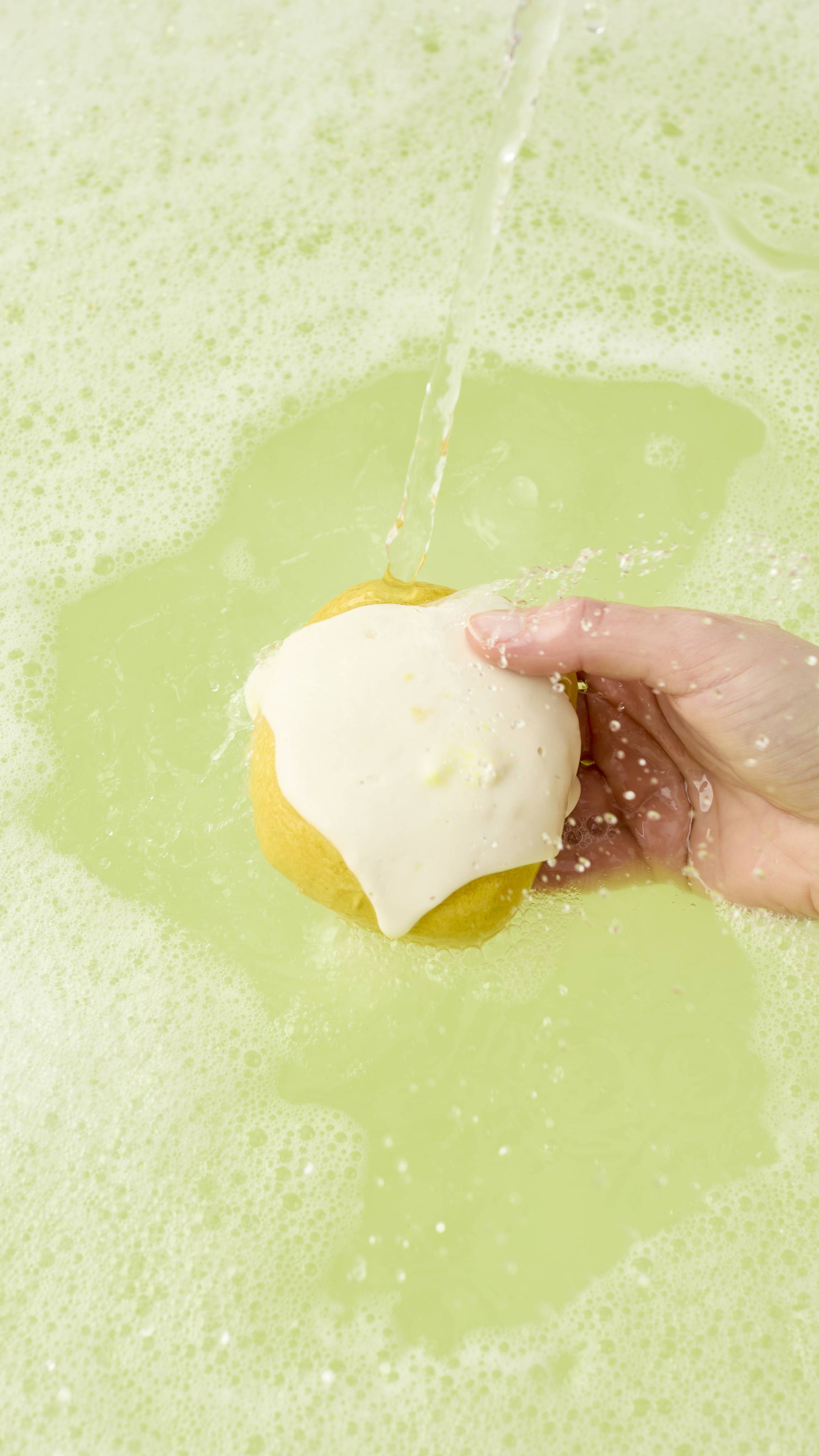 A model holds the "Easy Peasy Lemon Squeezy" bubble bar under a running tap. The water hits the white "icing", creating a frothy white lather that blends into the surrounding pale yellow, bubbly bathwater.