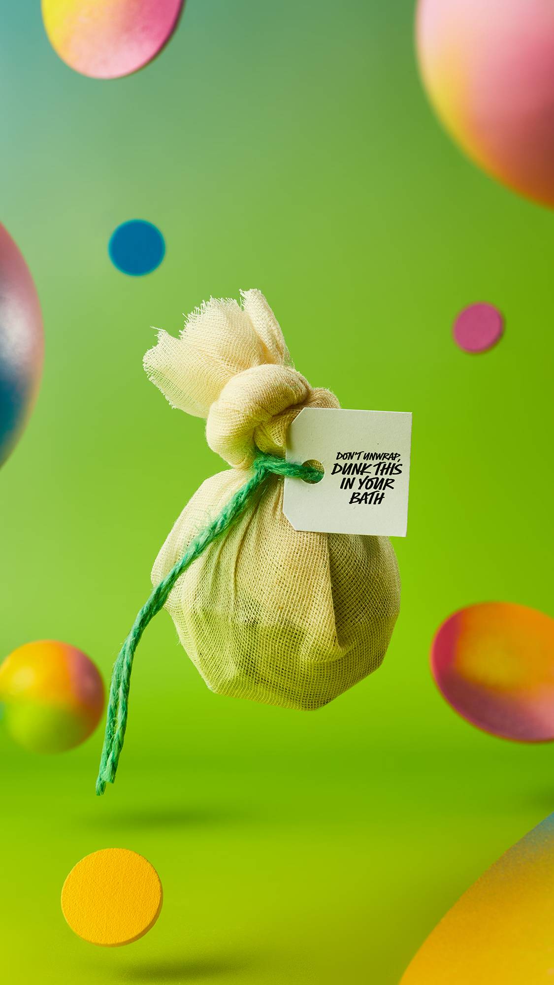 The image shows the muslin-wrapped, green Energising Matcha And Peppermint Cataplasm bath bomb suspended in mid-air tied with a green string on a green gradient background with colourful confetti falling around the edges.
