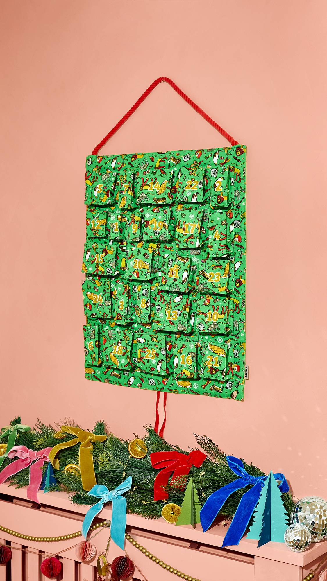 A green fabric Lush Advent calendar hanging by a red cord against a pink wall, featuring numbered pockets with colourful festive illustrations, positioned above a mantelpiece decorated with greenery and colourful bows.