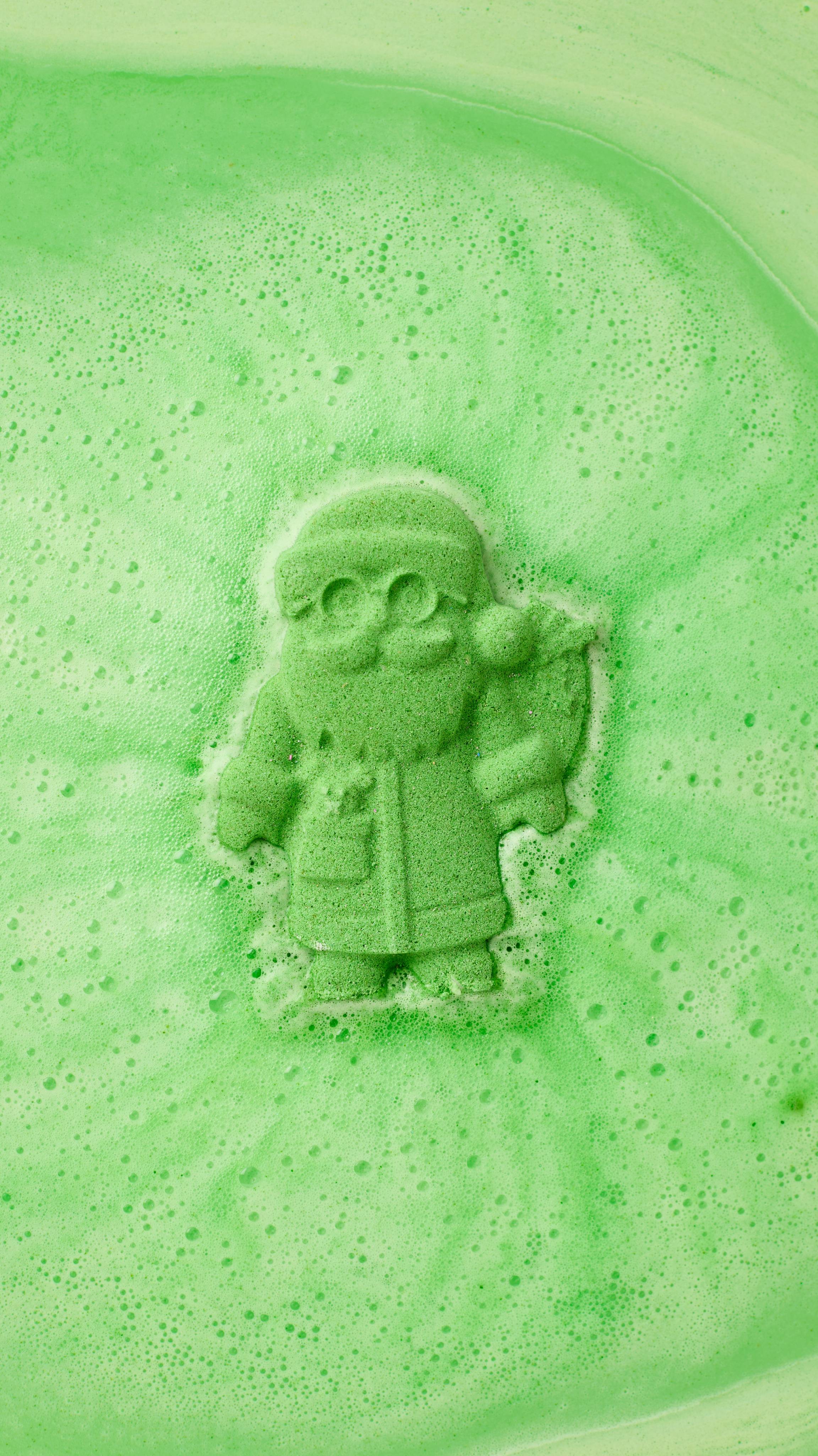 The image shows a speckled green Father Christmas bath bomb fizzing in bright green bath water. Abundant green foam and bubbles surround the Santa-shaped product as it dissolves.