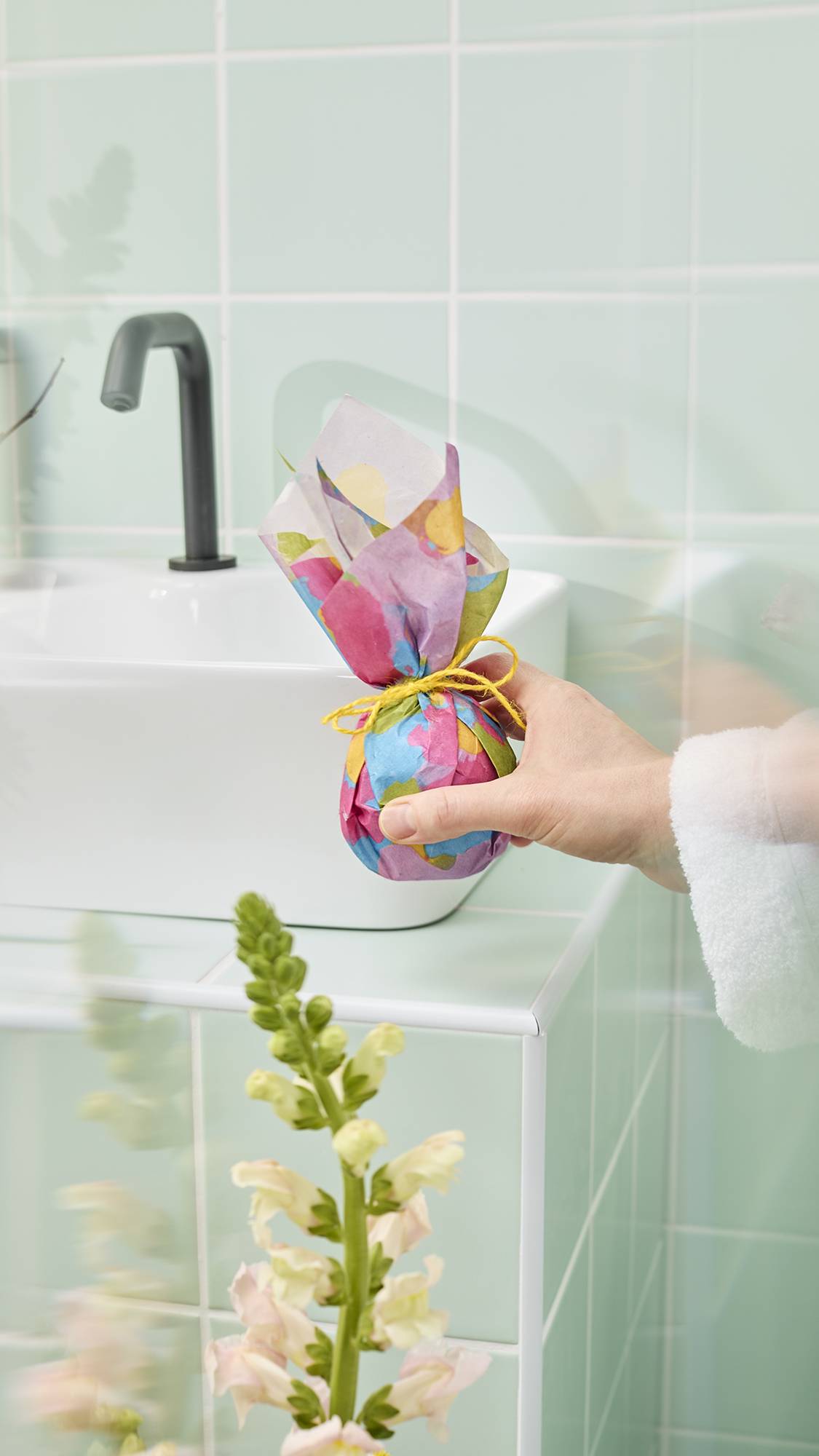 The image shows a hand holding a tied, "Flower Up" Lokta paper wrapped around a gift. The vibrant material, with hand-painted flowers in pink, purple, and blue on a lime-green background, is being secured over a gift on a pale green tiled surface.