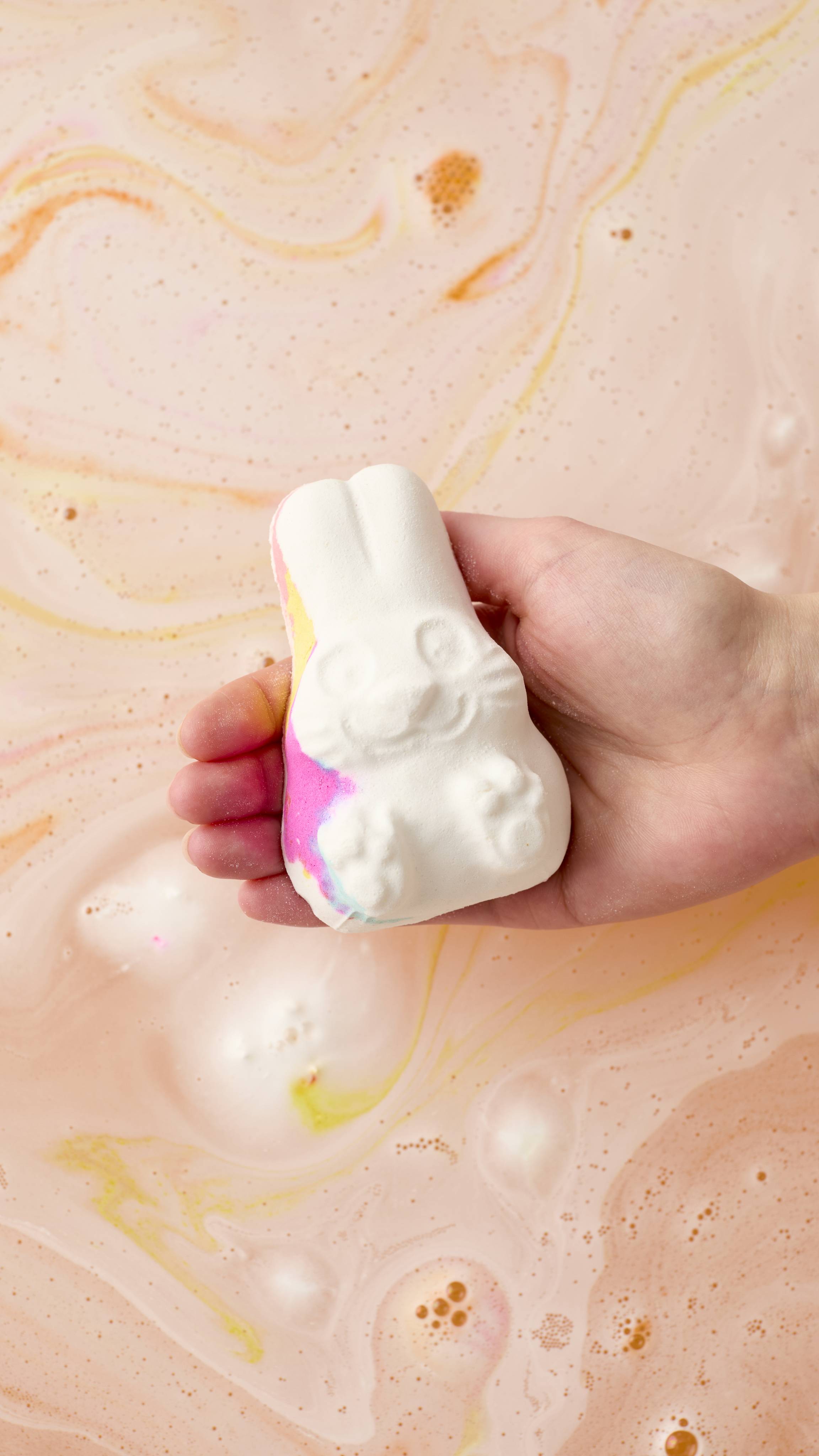 A hand holds a white, rabbit-shaped Follow The White Rabbit bath bomb over swirling peach and pink bathwater. The rabbit has colorful pink and yellow accents.
