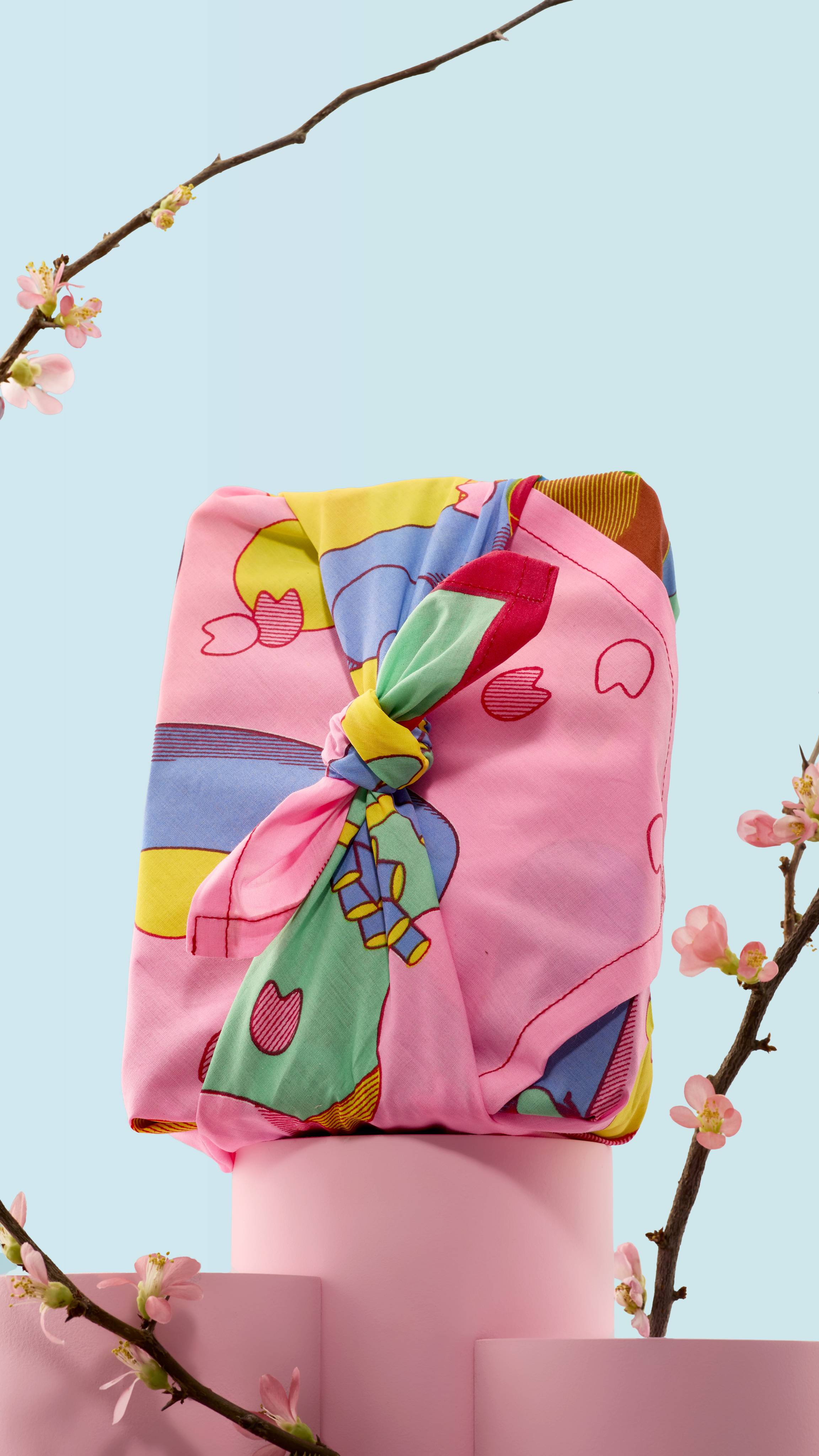 The image shows a gift box neatly wrapped in the vibrant "Forest Awaken" illustrated knot wrap and tied with a decorative knot. It sits on a pink podium, framed by delicate cherry blossom branches against a soft blue background. 