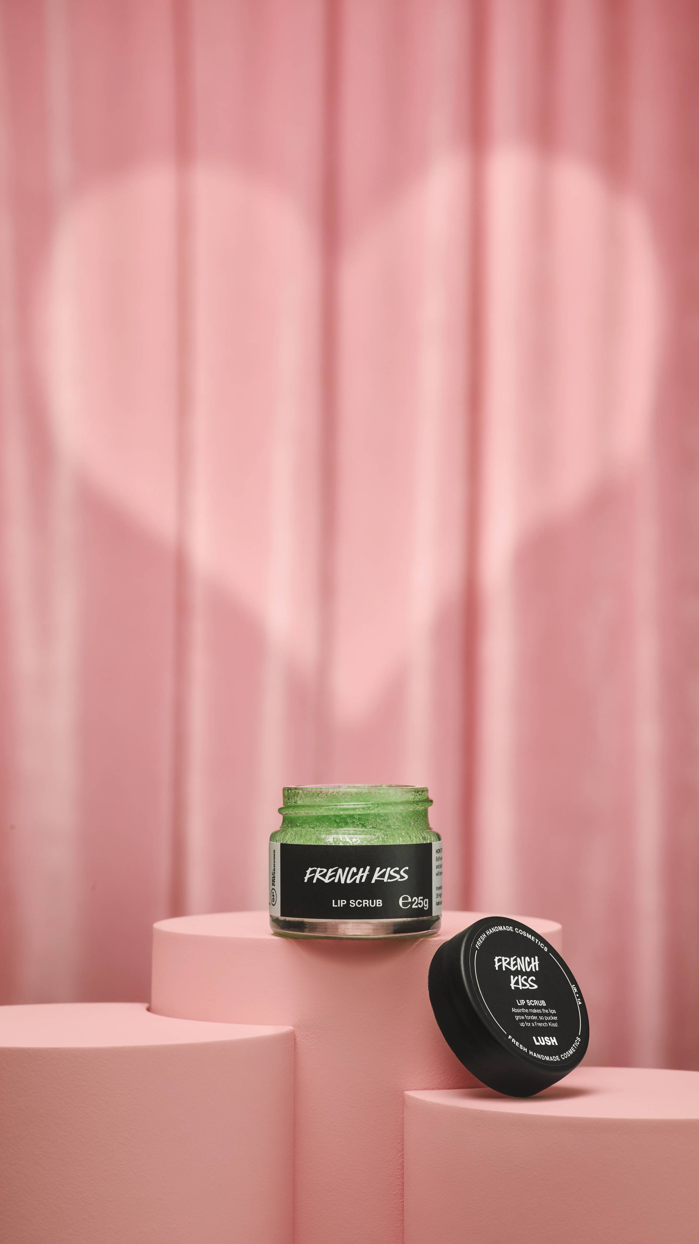 A small, clear glass jar of Lush "French Kiss" lip scrub sits on a pink podium. The lime-green scrub is shown with its black lid resting nearby against a pink curtain with a heart-shaped spotlight.